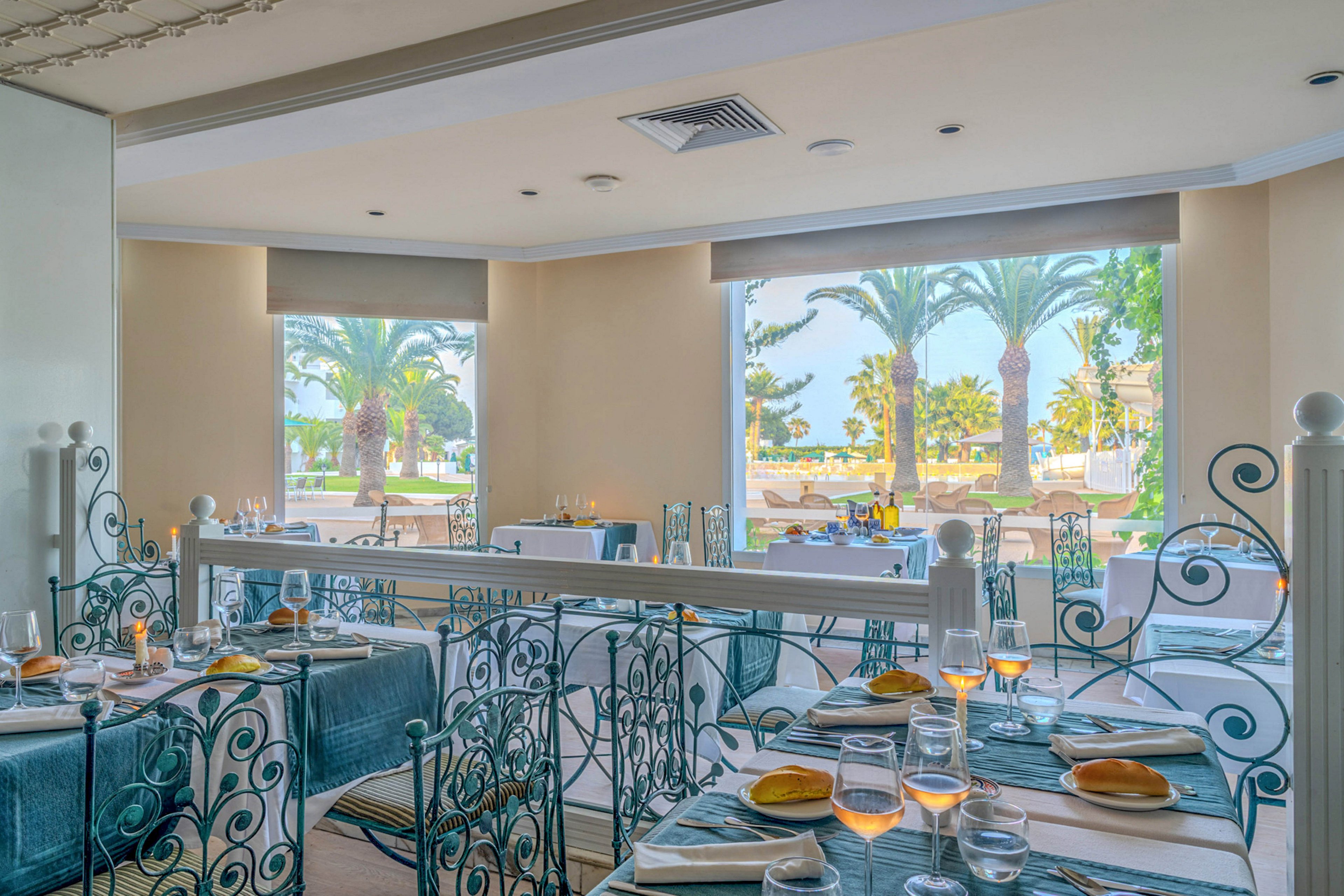 Bright, airy restaurant interior with neatly set tables overlooks a sunny outdoor terrace lined with palm trees. Tables are dressed in blue and white linens, with bread rolls, glassware, and candles creating a refined dining atmosphere. Large windows frame the coastal landscape, allowing natural light to fill the space. Ornate metal railings and soft colors contribute to a relaxed yet upscale seaside mood.
