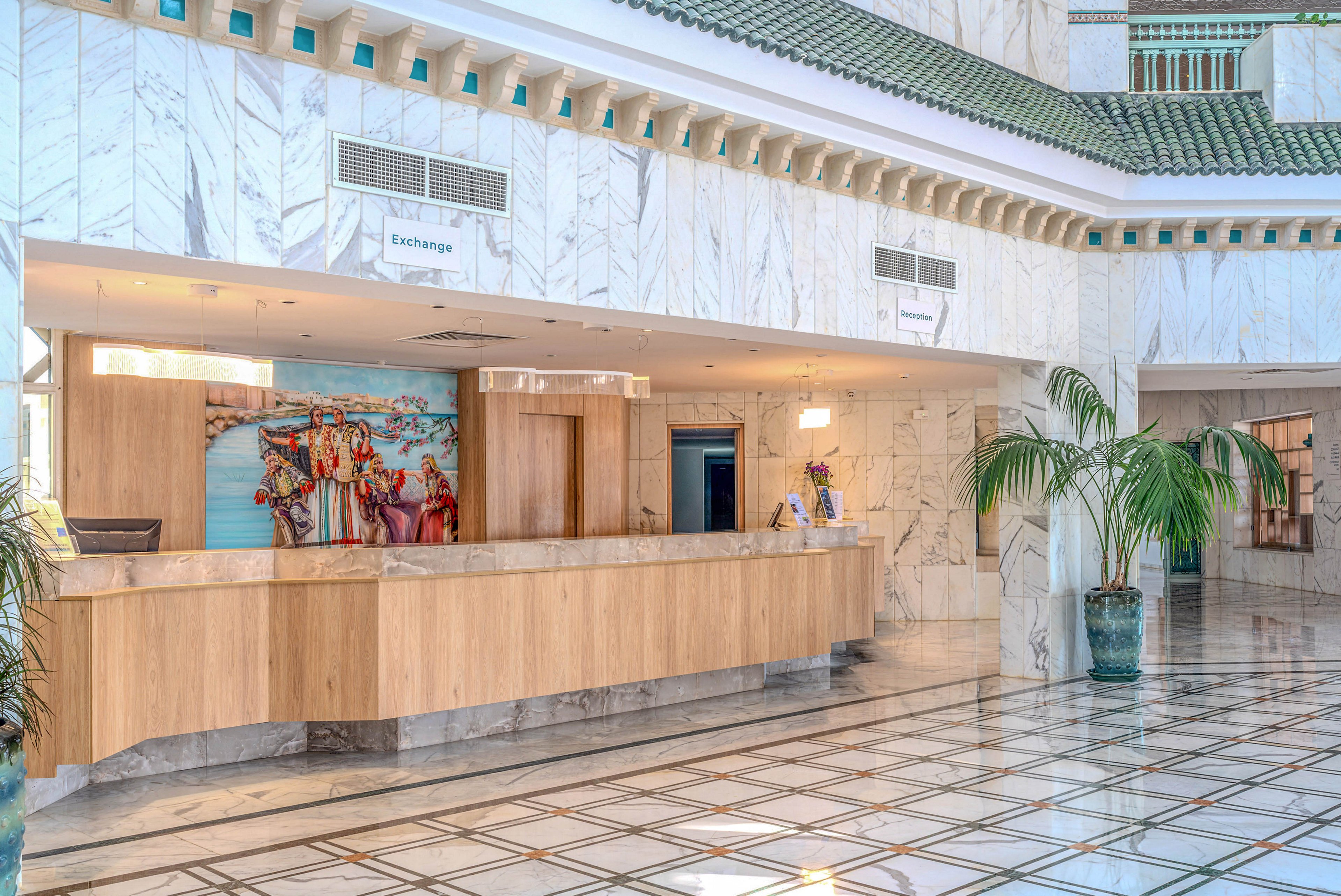 A spacious hotel lobby features a long wooden reception desk set against white marble walls. The floor is tiled with a glossy geometric pattern that reflects the light from overhead fixtures. A colorful mural decorates the wall behind the desk, adding contrast to the otherwise neutral interior. Large potted palm plants frame the area, contributing to a relaxed, resort-like atmosphere.