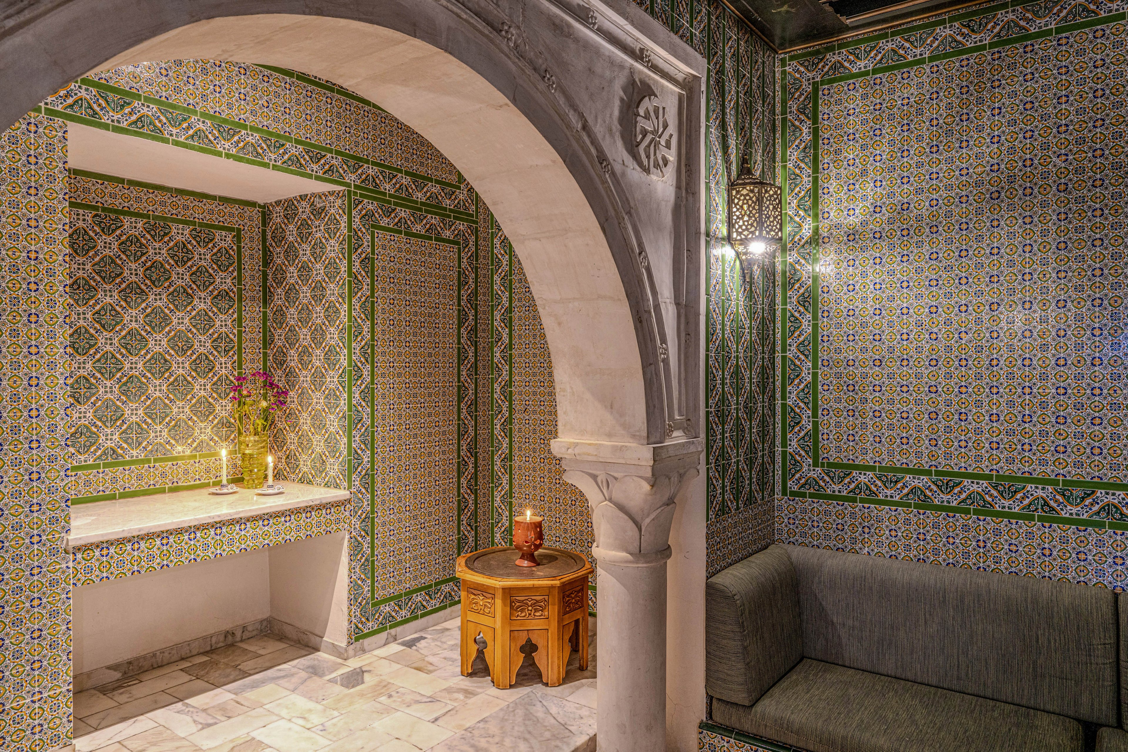 An ornate interior lounge features intricate geometric tilework covering the walls and archway. A carved stone arch frames a cozy seating area with a cushioned bench and a small wooden side table holding a lit candle. Decorative lantern lighting and a vase of flowers on a tiled niche add warmth and detail to the space. The overall ambiance is intimate, traditional, and richly patterned.