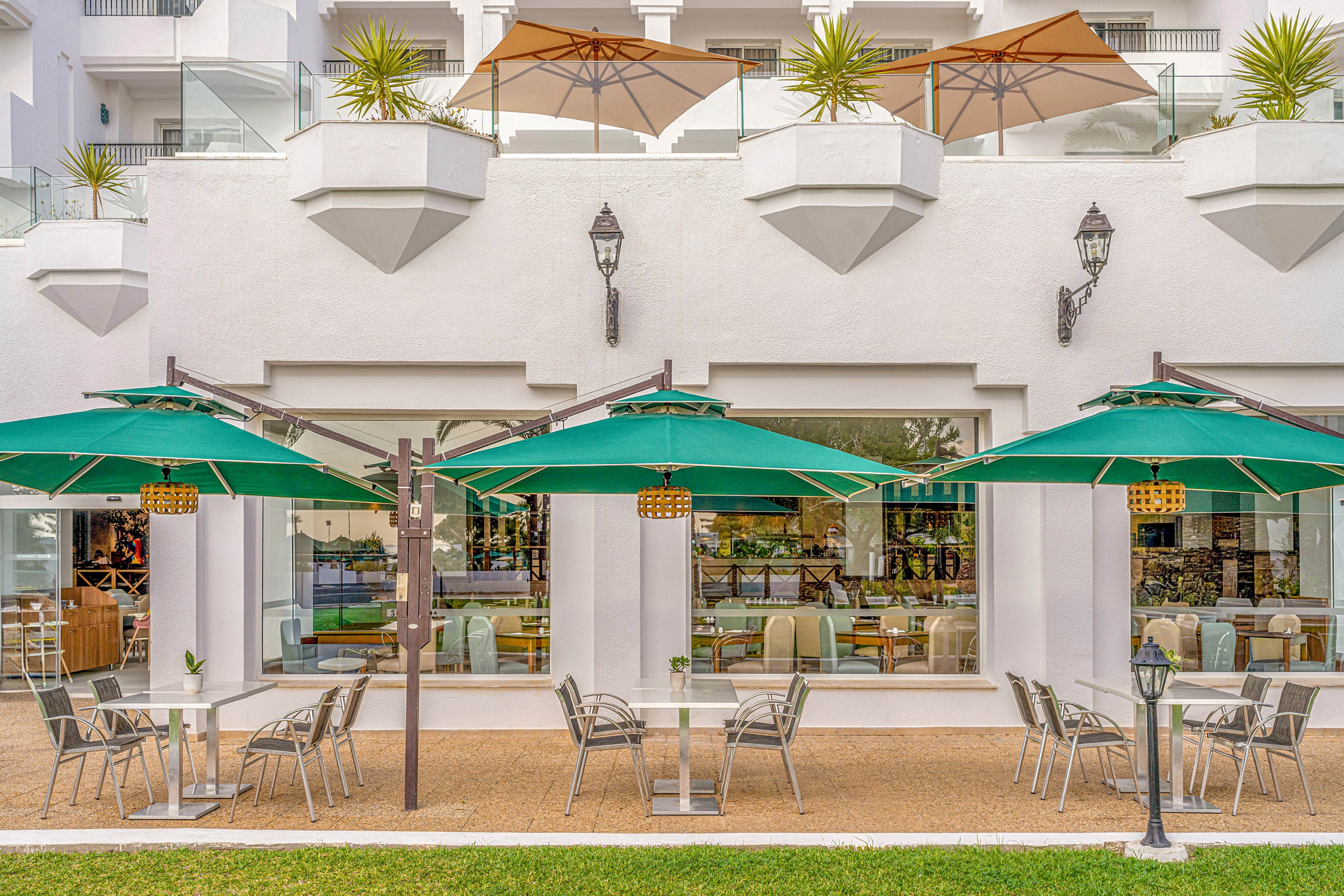 An outdoor café terrace is set against a white resort building with large windows and decorative wall lanterns. Green patio umbrellas shade metal tables and chairs arranged on a paved area beside a strip of green grass. Above, beige umbrellas and potted plants line the upper level balconies, adding height and texture. The scene suggests a relaxed vacation atmosphere with comfortable seating for dining or drinks.