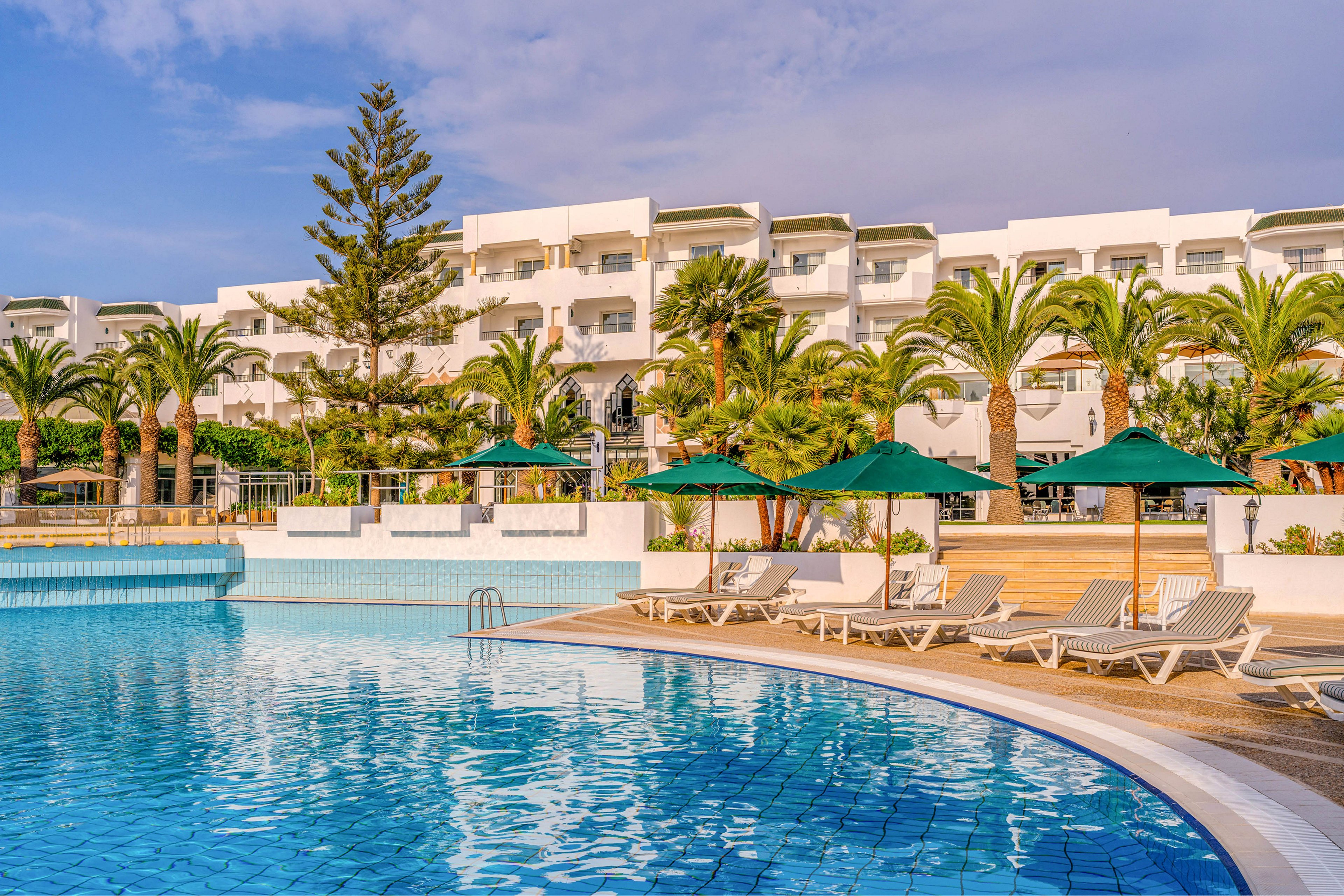 A large outdoor swimming pool is surrounded by white sun loungers and green parasols at a coastal resort. Palm trees and lush greenery frame the pool area, adding a tropical feel to the scene. In the background, a white multi-story hotel building with balconies overlooks the water. The bright blue sky and warm sunlight create a relaxed vacation atmosphere.