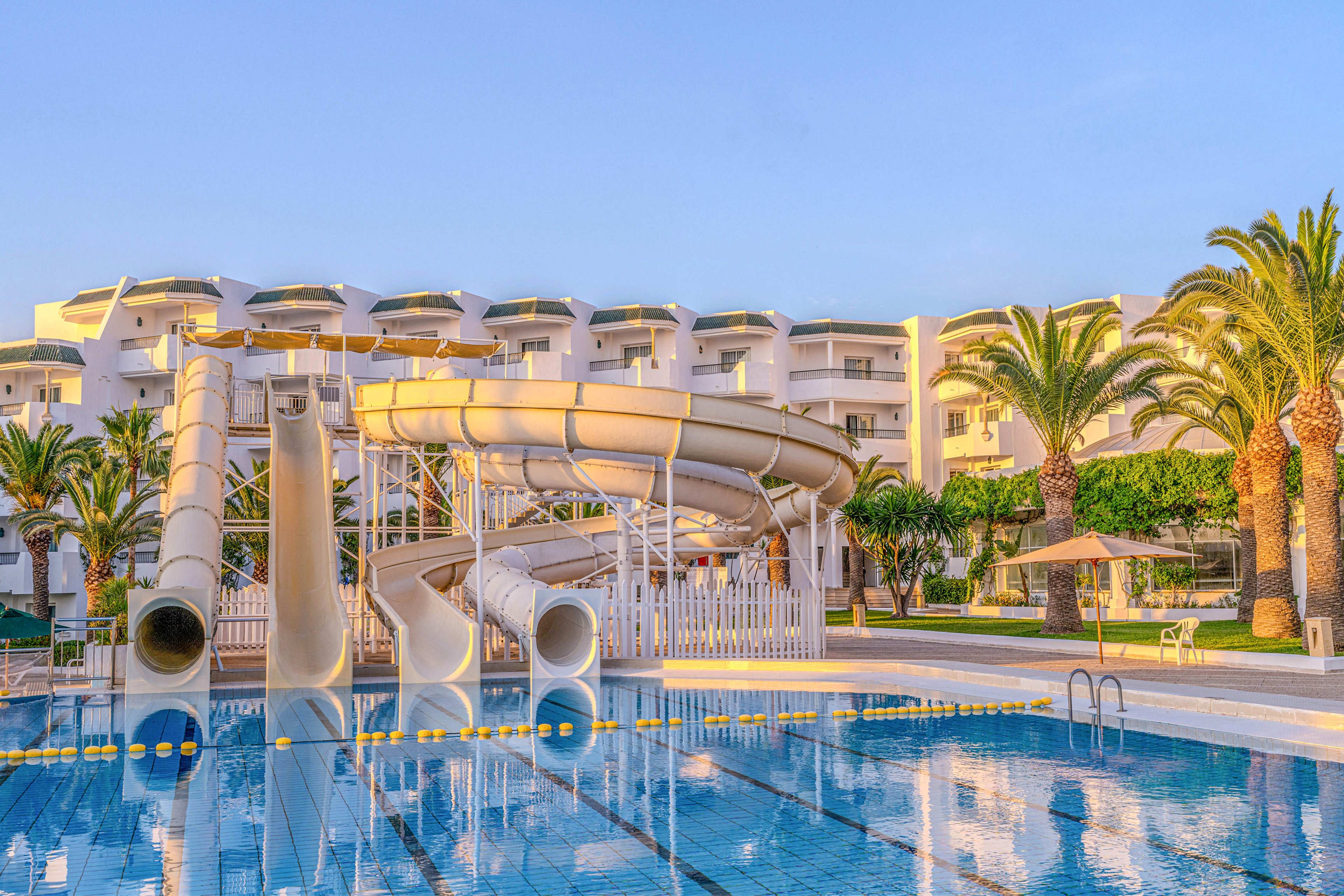 Large white waterslides curve above a clear blue swimming pool at a modern resort. The multi-story hotel building with balconies forms the backdrop, reflecting warm sunset or sunrise light. Tall palm trees and neatly trimmed greenery line the poolside, adding a tropical atmosphere. Pool ladders, railings, and shaded seating areas suggest a relaxing vacation setting with family-friendly water activities.
