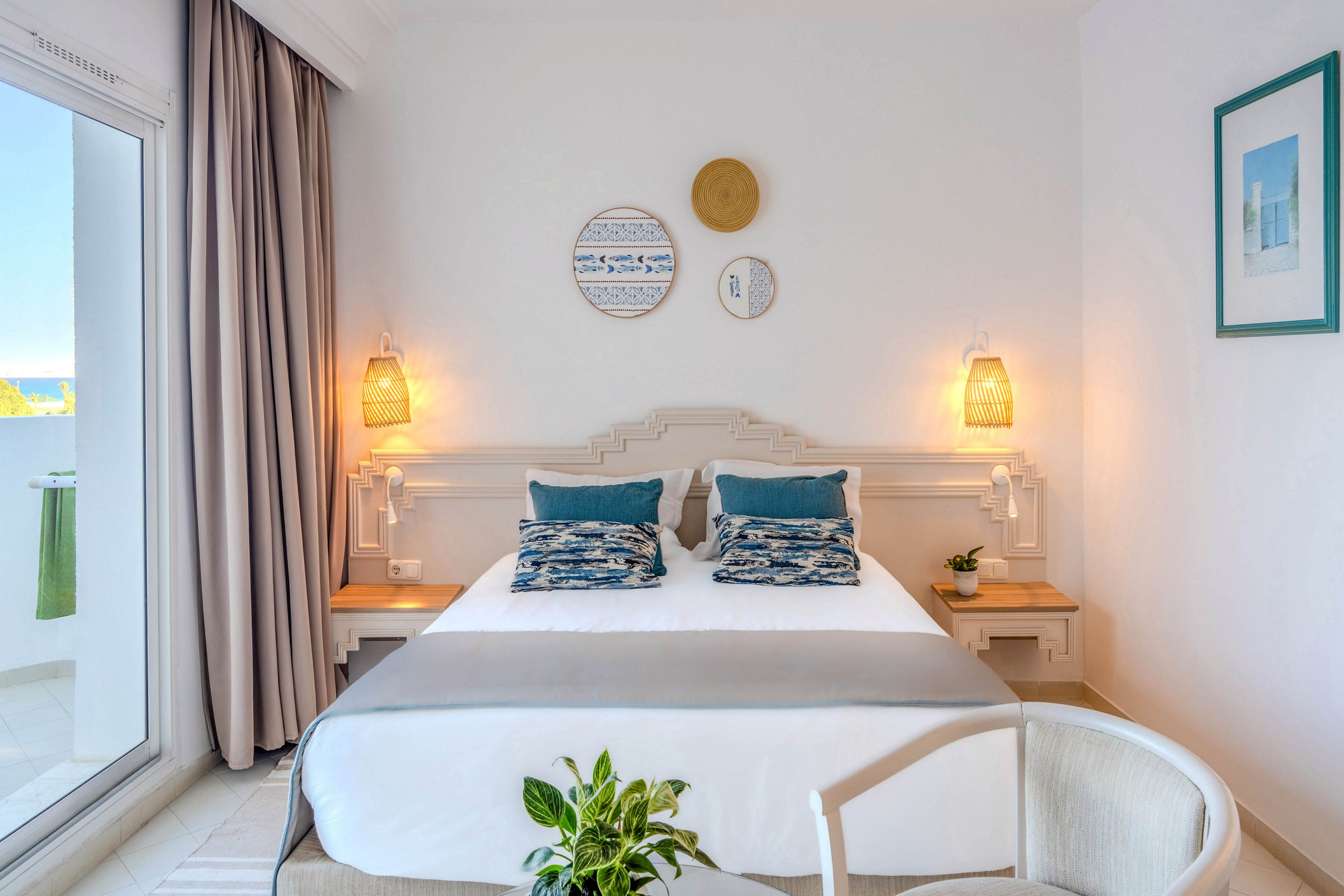 A neatly made double bed with white linens and blue accent pillows is centered in a bright, modern bedroom. Two warm wall sconces flank the bed above light wooden nightstands, adding a cozy glow. Decorative round wall art and a framed picture enhance the coastal style, while a potted plant and soft armchair sit in the foreground. A large window with curtains opens to a sunny outdoor view, filling the room with natural light.