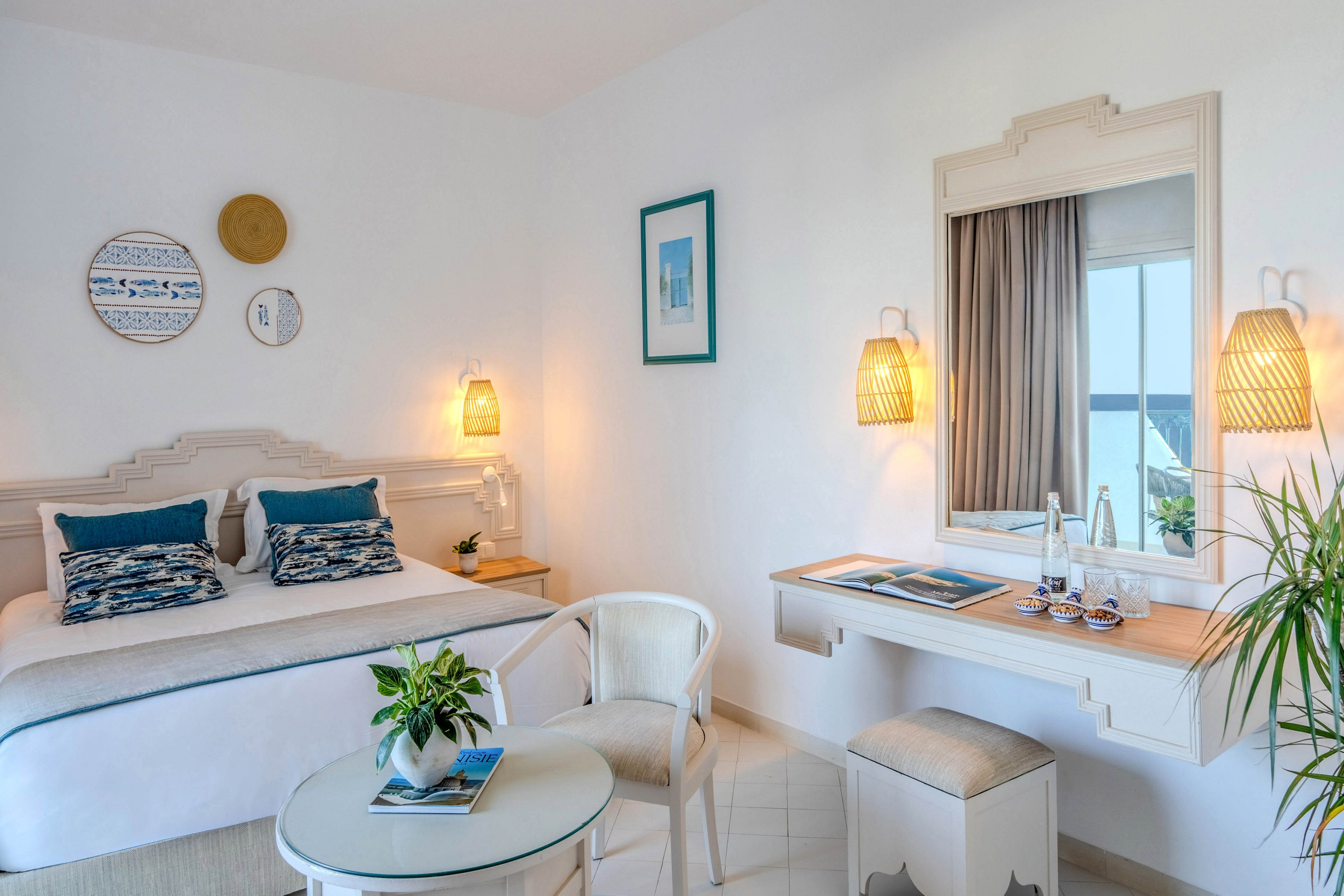 A light and airy hotel bedroom features a neatly made double bed with blue accent pillows and patterned cushions. The room includes a small round table with chairs, a vanity desk with a mirror, and warm woven wall lamps. Decorative wall art and a potted plant add a relaxed coastal touch, while a window with curtains reveals a view of the sea. The overall palette is soft white with blue and natural wood tones, creating a calm vacation atmosphere.