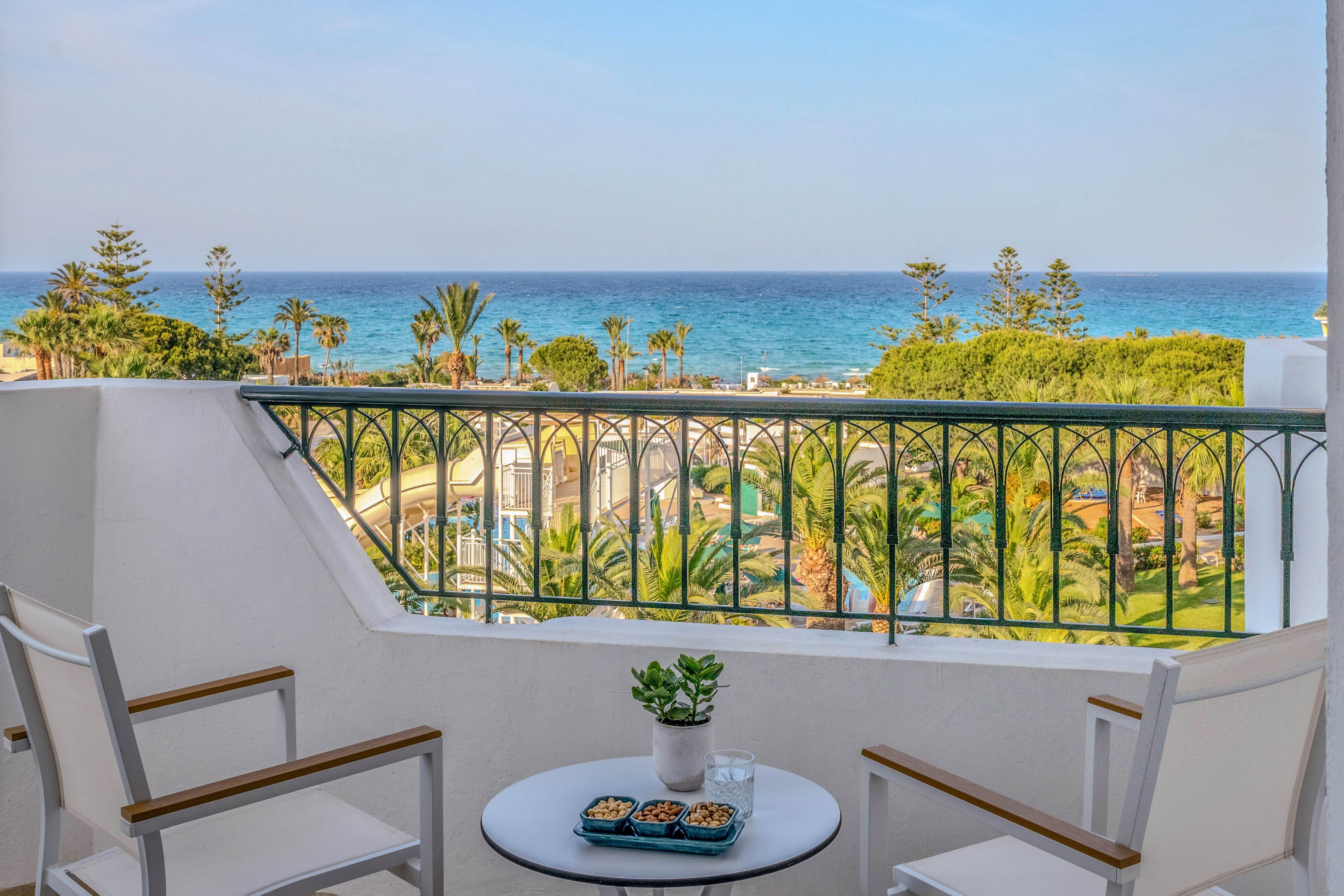A small balcony with white chairs and a round table overlooks a lush coastal resort. On the table are two glasses of water, a small potted plant, and a tray of snacks, suggesting a relaxed outdoor break. Beyond the decorative railing, palm trees and gardens lead down to the vivid blue sea under a clear sky. The scene conveys a tranquil vacation atmosphere with bright, natural colors.