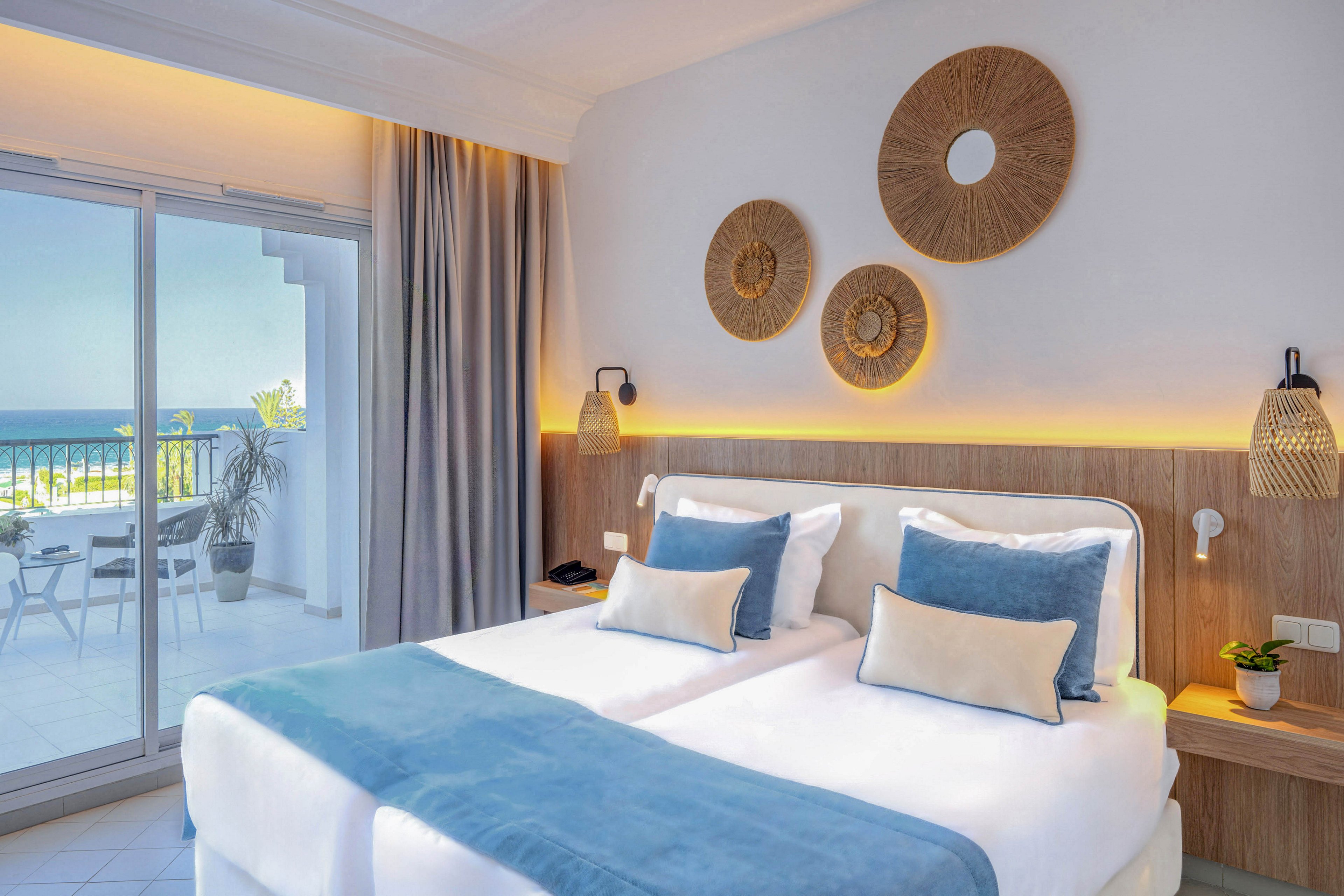 Modern twin beds with white linens and light blue throws sit in a bright hotel bedroom. A sliding glass door opens to a balcony with chairs and a view of the sea. Warm accent lighting highlights woven wall decor and bedside lamps, creating a cozy atmosphere. Neutral walls and curtains complement the soft blue palette for a relaxing coastal feel.