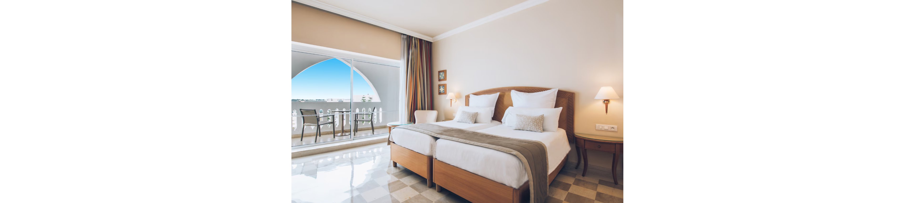 A spacious hotel bedroom features two neatly made twin beds with white linens and neutral-toned throws. The room opens onto a balcony through large sliding glass doors, revealing a clear blue sky and distant water view. Soft wall sconces and a bedside lamp create warm ambient lighting against beige walls. A small seating area on the balcony invites guests to relax and enjoy the coastal scenery.
