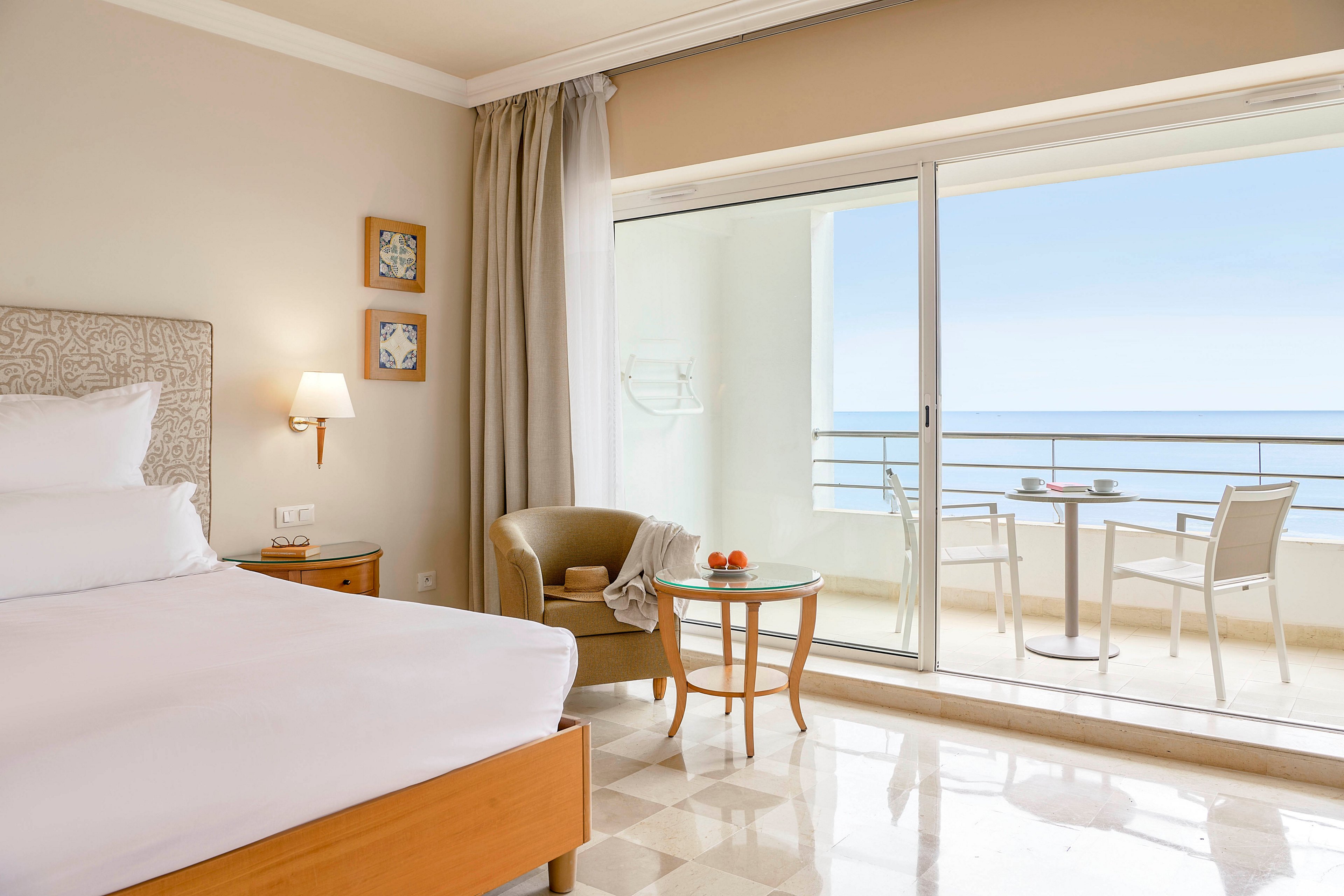 A modern hotel room features a neatly made bed with white linens and a light wood frame. Beside the bed, a small armchair and round glass table hold a few pieces of fruit, adding a touch of color. Large sliding glass doors open onto a balcony with a table, chairs, and a clear view of the calm sea and blue sky. The interior decor is minimal and airy, with neutral tones and soft natural light creating a relaxing atmosphere.