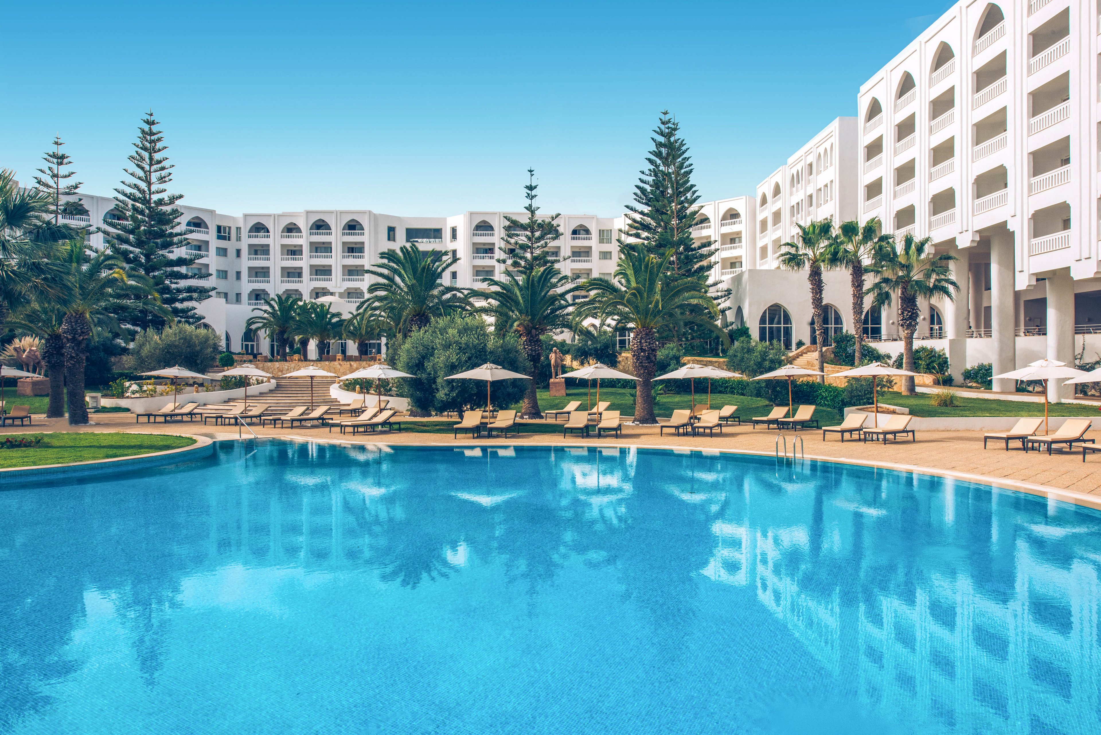 A large, clear blue swimming pool dominates the foreground of a luxury resort hotel. White multi-story buildings with balconies curve around the pool area, framed by tall palm trees and lush greenery. Sun loungers and umbrellas line the poolside terraces, suggesting a relaxing vacation atmosphere. The bright blue sky and calm water create a serene, sunny holiday setting.