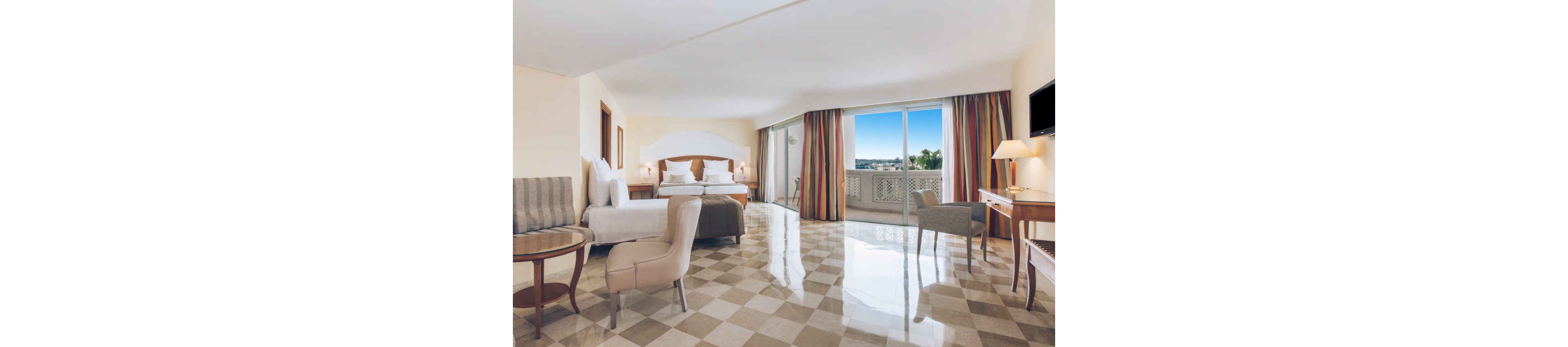 Spacious hotel suite featuring a large bed, seating area, and writing desk. The room is decorated in neutral tones with colorful floor-to-ceiling curtains framing a wide balcony door. Sunlight reflects off the polished checkered floor, creating a bright and airy atmosphere. A flat-screen TV, table lamp, and outdoor view complete the modern, comfortable setting.