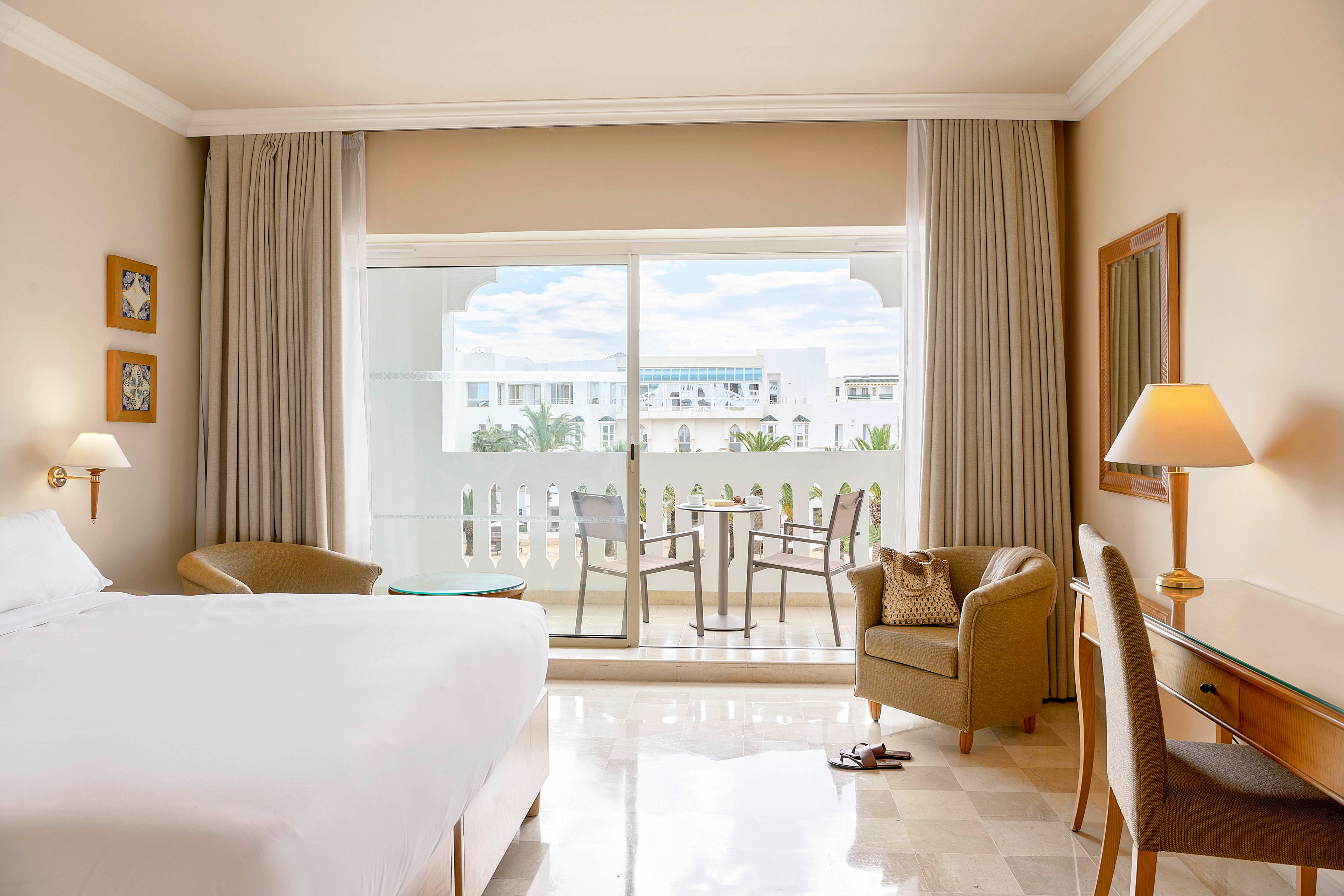 A spacious hotel room features a large neatly made bed and neutral-toned decor. Floor-to-ceiling curtains frame a sliding glass door that opens onto a balcony with two chairs and a small table. Soft lighting from bedside and desk lamps creates a warm, inviting atmosphere. Outside, white buildings and palm trees are visible under a partly cloudy sky, suggesting a resort or coastal location.