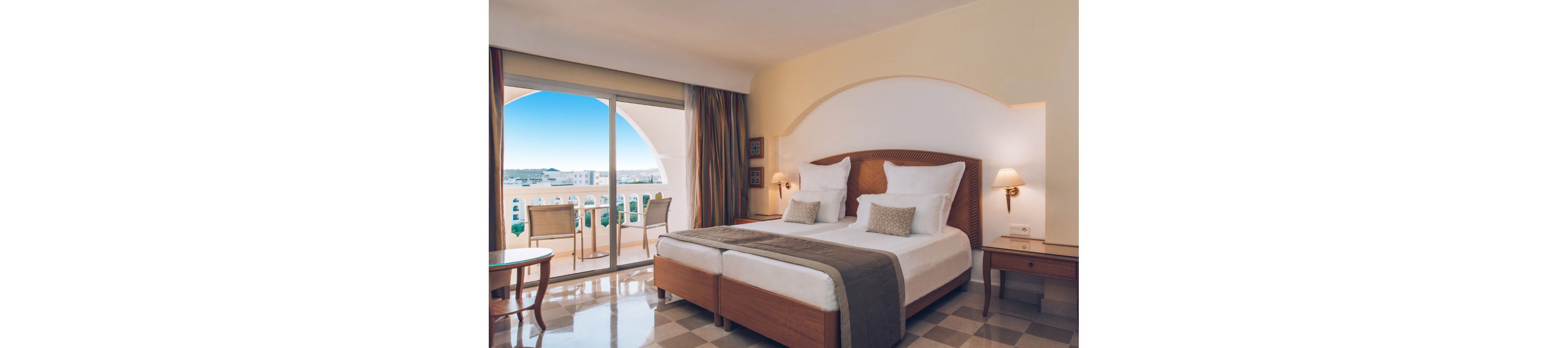 A neatly arranged hotel bedroom features two single beds pushed together with crisp white linens and accent pillows. The room includes wooden bedside tables, wall-mounted lamps, and a desk area, creating a comfortable and functional space. Large sliding glass doors open onto a balcony with chairs and a table, overlooking white buildings and a clear blue sky. Warm neutral tones and natural light give the interior a calm, inviting atmosphere.