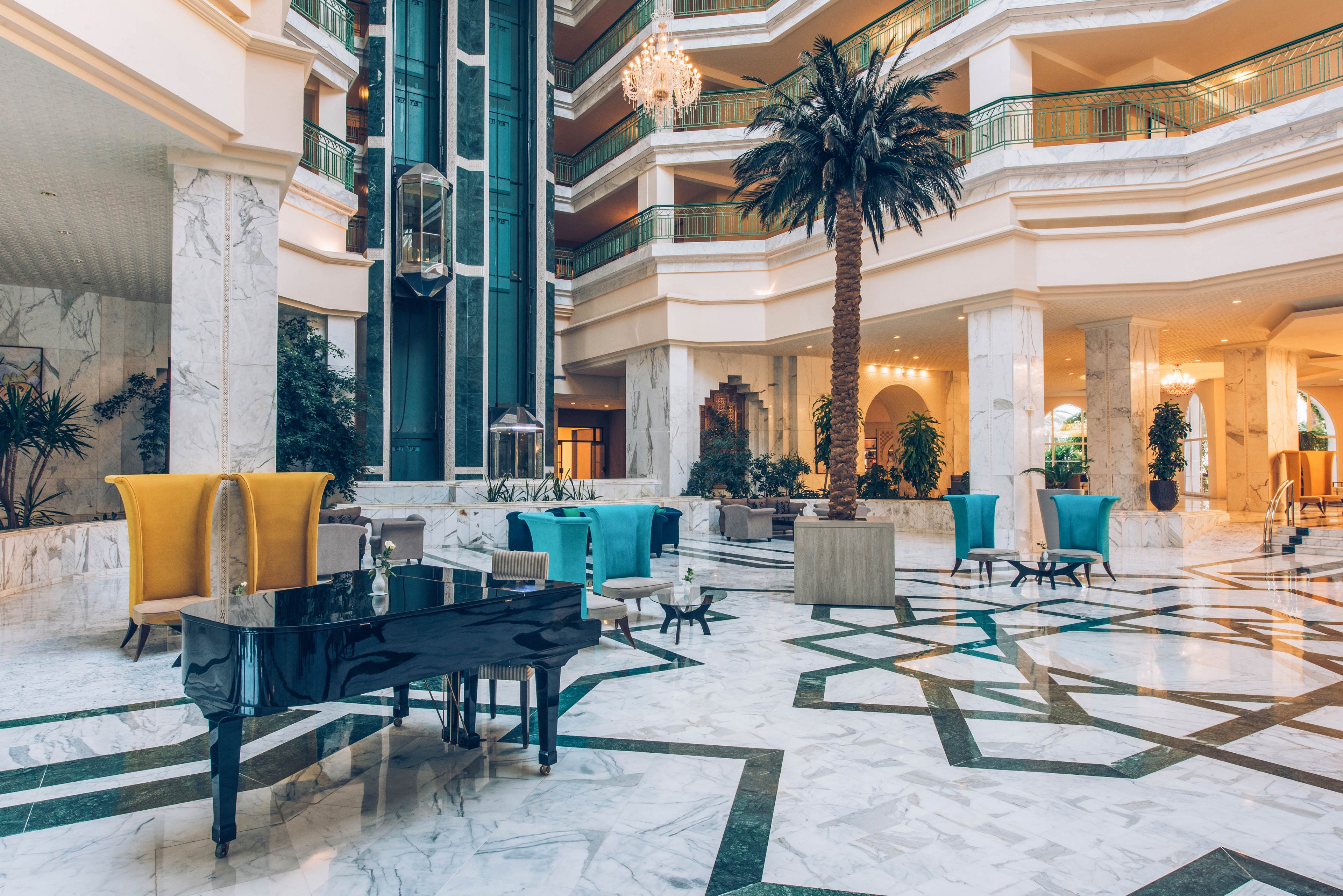 A spacious hotel lobby features a glossy black grand piano set on a polished marble floor. Colorful modern armchairs and small tables are arranged in seating areas around the open atrium. A tall indoor palm tree and glass elevator add vertical interest to the multi-level interior. Warm ambient lighting and decorative chandeliers create an elegant, welcoming atmosphere.