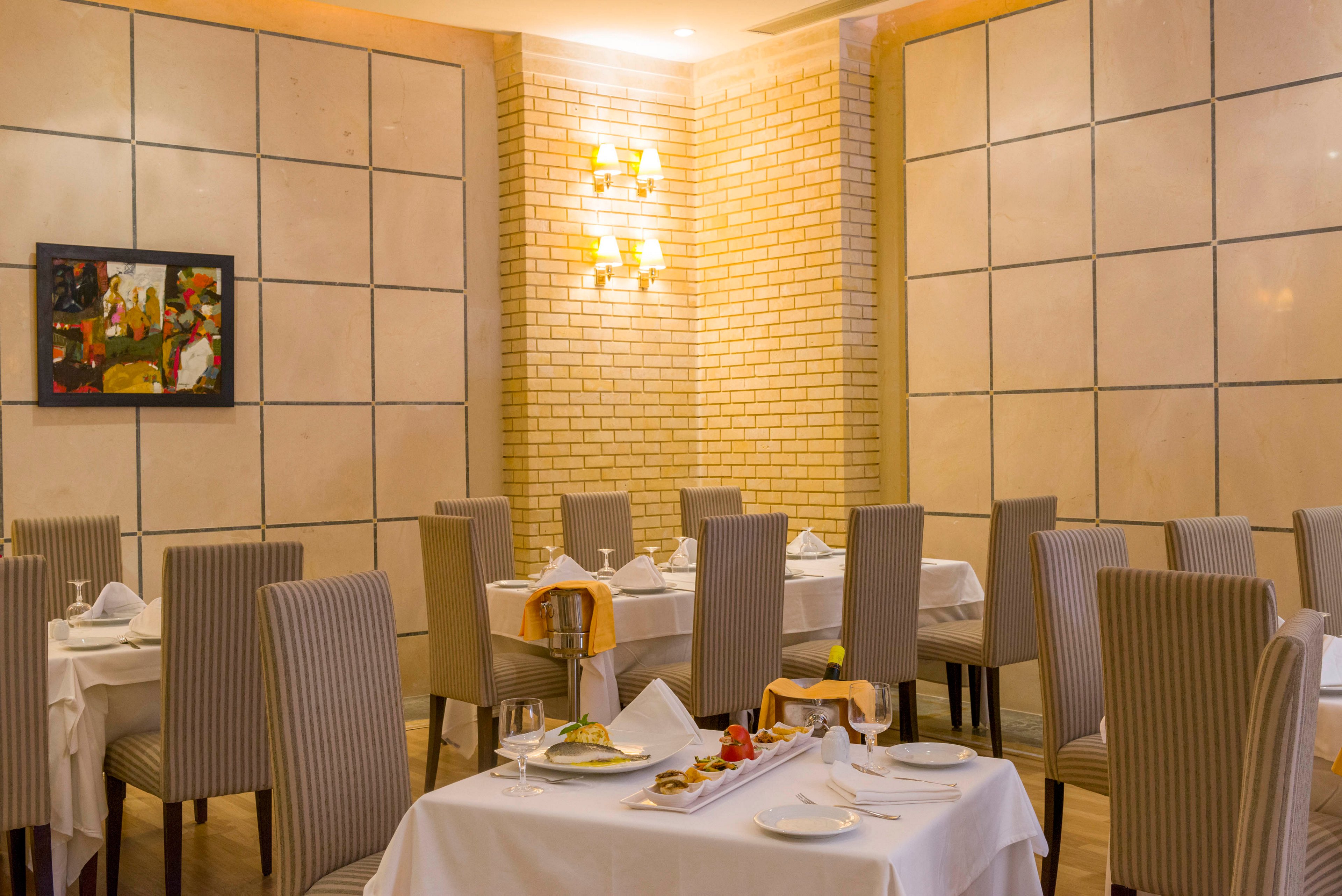 An upscale restaurant dining room features neatly arranged tables covered with white tablecloths. Tall, striped upholstered chairs surround each table, which is set with plates, glasses, and folded napkins. Warm wall sconces illuminate the light brick and paneled walls, creating a cozy yet refined atmosphere. A colorful abstract painting adds a touch of modern art to the otherwise neutral interior.