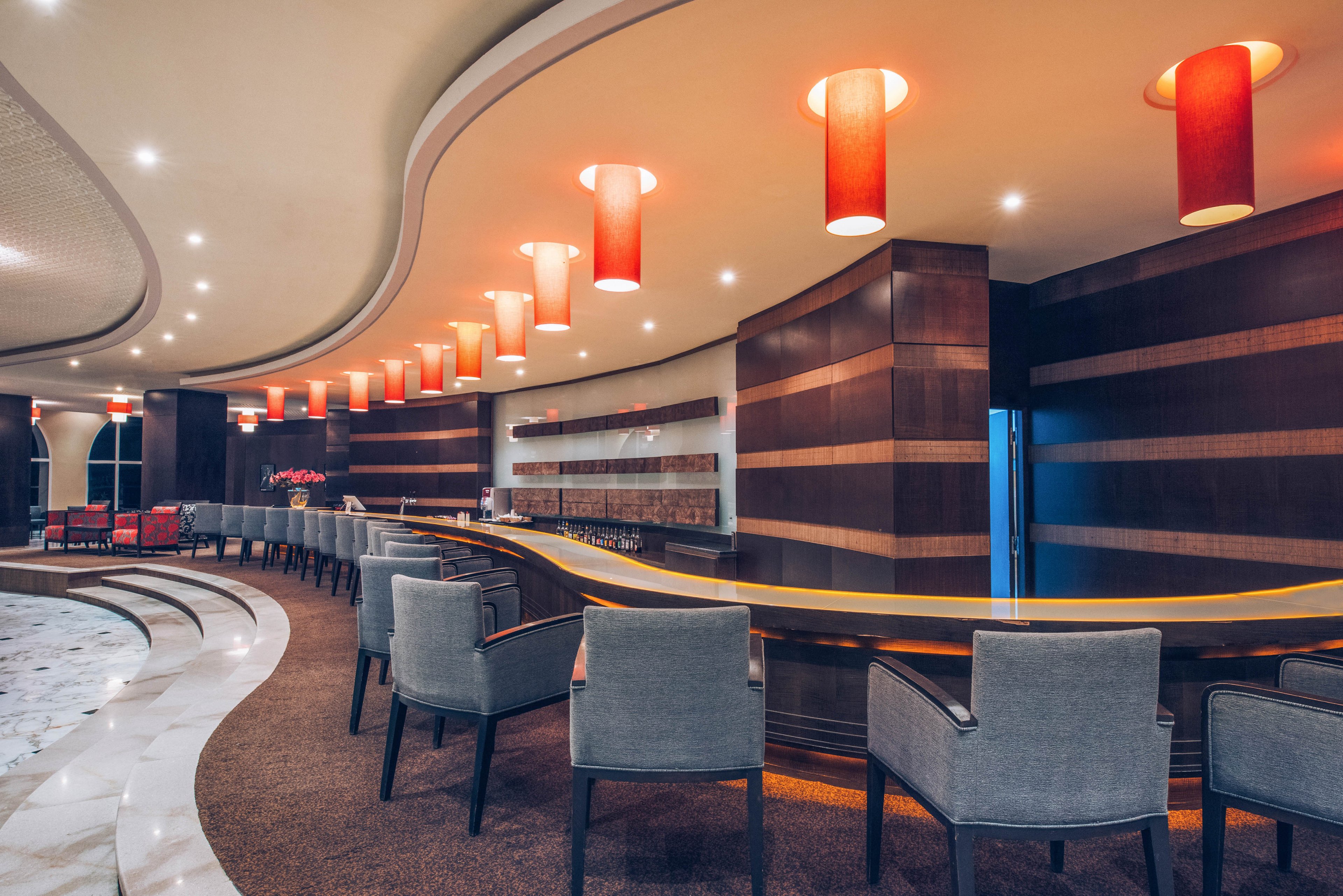 Contemporary bar interior featuring a long curved counter lined with upholstered chairs. Warm wooden paneling and striped wall details create a sleek, upscale atmosphere. Red cylindrical pendant lights hang from the ceiling, casting a soft glow over the bar area. The space appears empty, emphasizing the clean lines, polished surfaces, and modern design elements.
