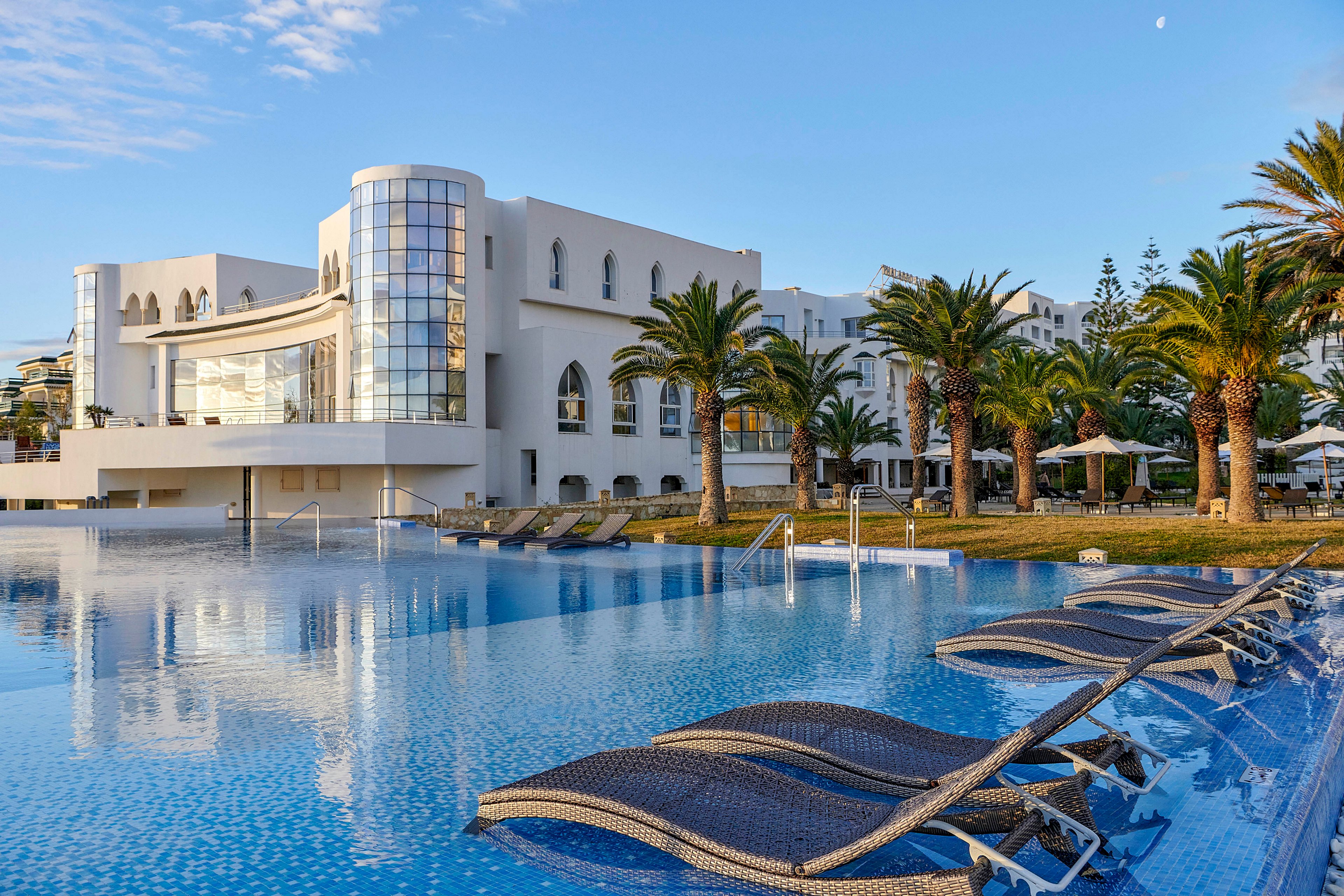 A modern white resort building overlooks a large, tranquil infinity pool. Wicker lounge chairs are partially submerged in the shallow water, inviting guests to relax by the poolside. Palm trees and neatly kept lawns frame the outdoor area, suggesting a warm coastal or tropical destination. The soft evening light and clear sky create a serene and upscale vacation atmosphere.