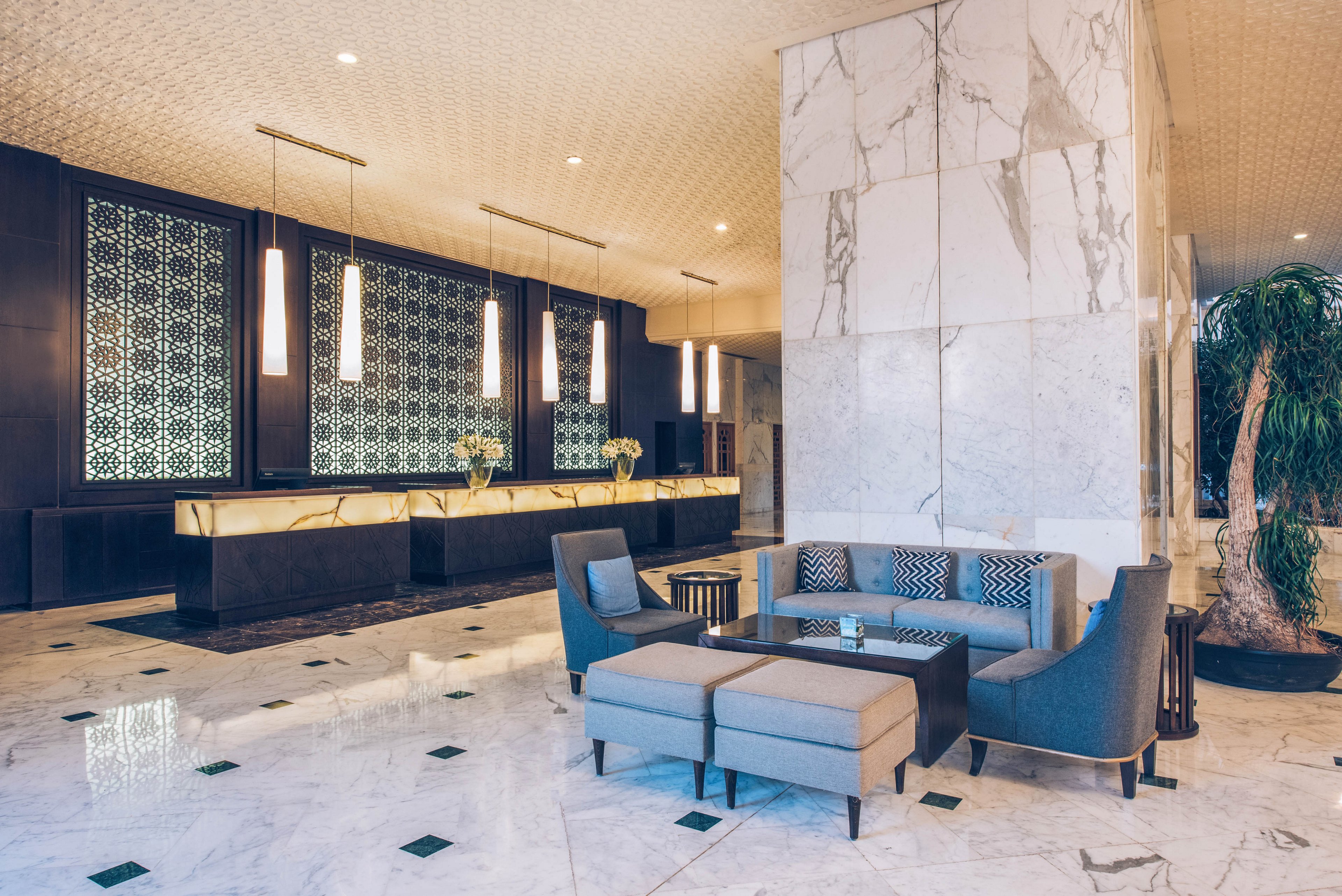 A contemporary hotel lobby features a sleek marble reception desk and patterned backlit wall panels. In the foreground, a seating area with gray sofas, armchairs, and a low coffee table sits on a polished marble floor. Tall pendant lights hang above the front desk, casting a warm glow across the space. A large indoor plant adds a touch of greenery to the otherwise minimalist, upscale interior.