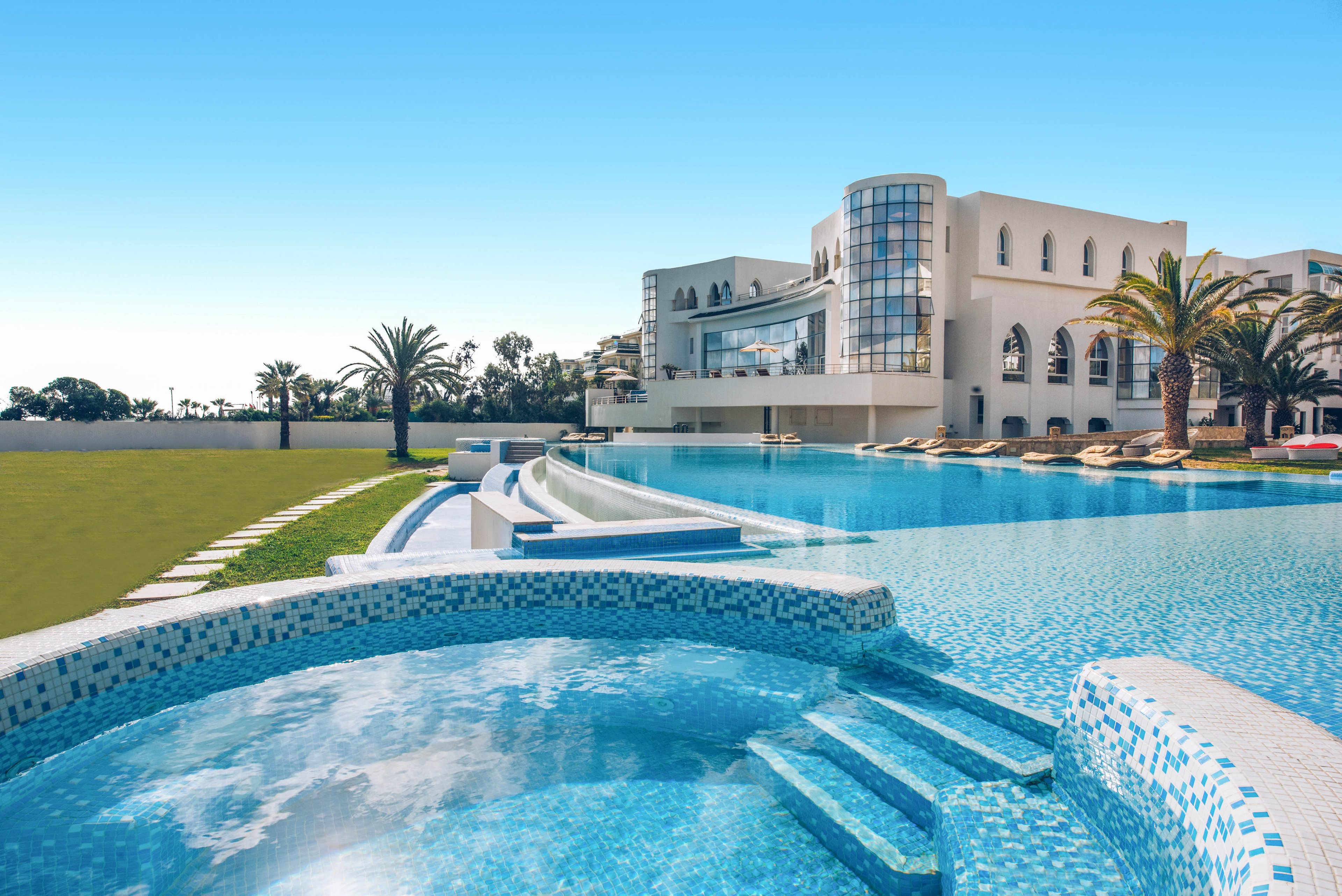A large luxury resort features an expansive infinity pool in the foreground with mosaic-tiled steps leading into the water. The modern white building has curved glass windows and multiple levels overlooking the pool area. Sun loungers and palm trees line the poolside, suggesting a relaxing vacation atmosphere. The sky is clear and blue, enhancing the bright, tranquil setting.