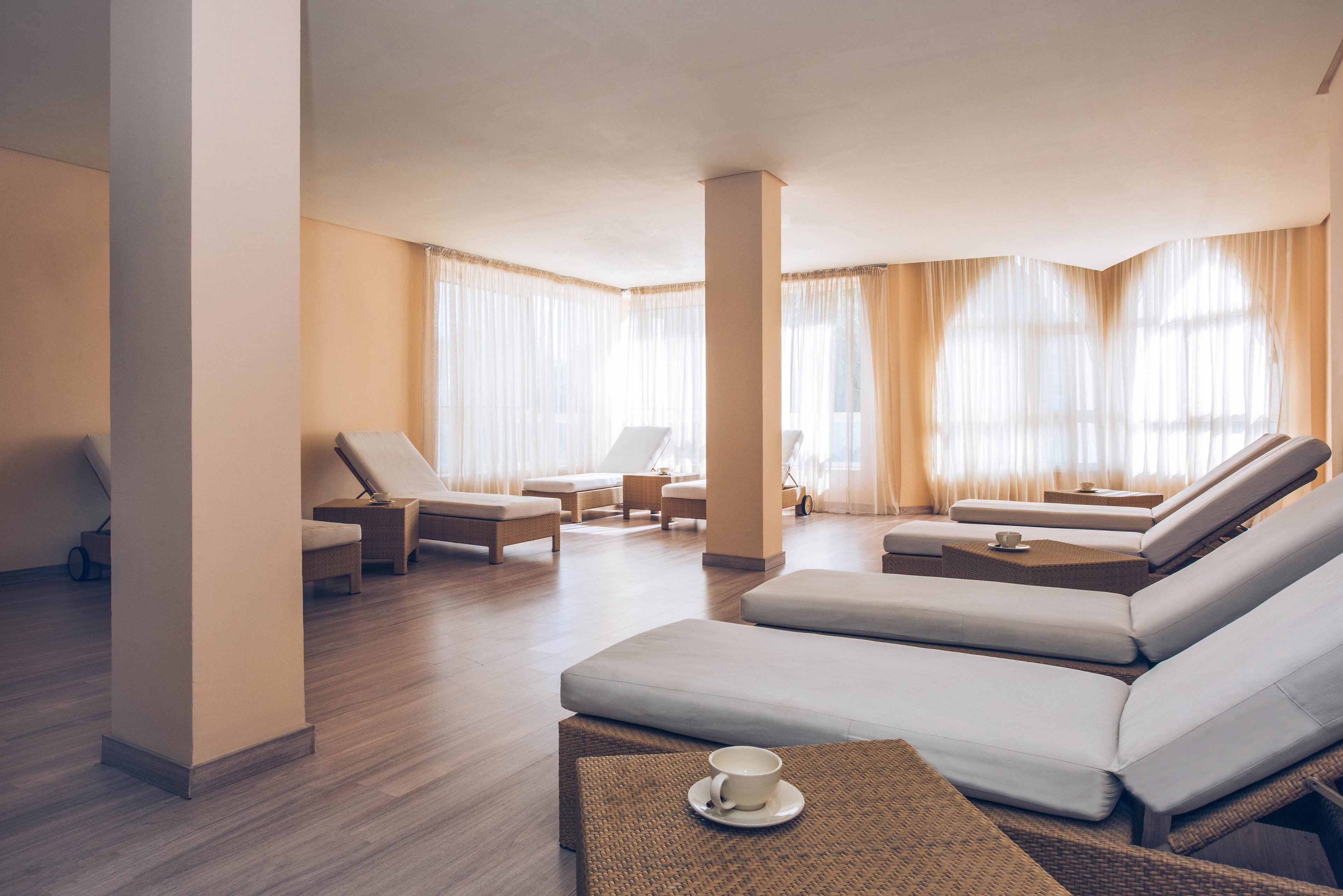 A serene spa lounge features several cushioned wicker loungers arranged in a spacious, sunlit room. Soft natural light filters through sheer curtains covering large corner windows. Neutral beige and cream tones create a calm, minimalist atmosphere. A small table with a cup and saucer in the foreground adds a touch of comfort and hospitality.