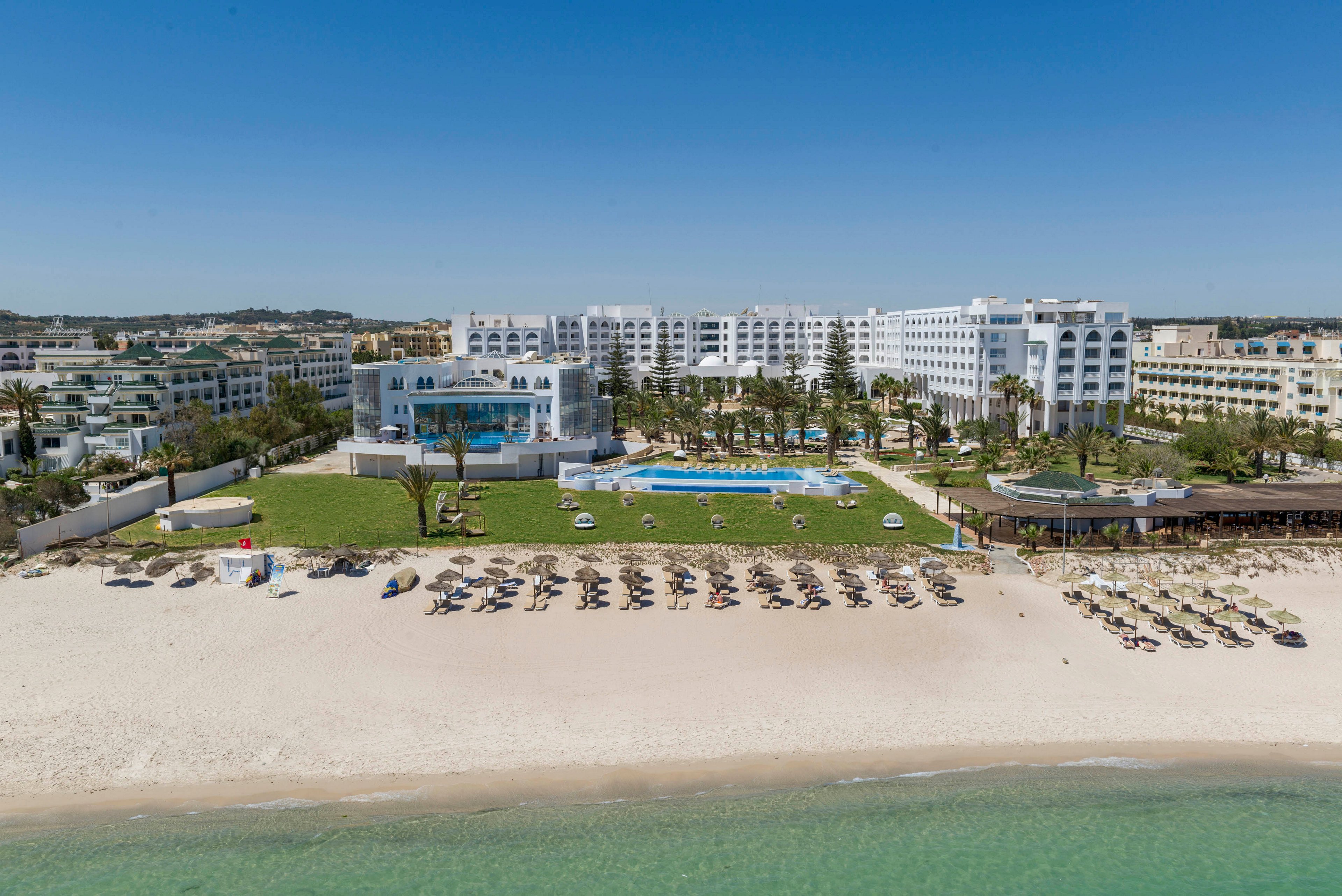 A large white beachfront resort overlooks a wide sandy beach lined with rows of sun loungers and umbrellas. In front of the hotel buildings, a green lawn surrounds an outdoor swimming pool and terrace area. The calm turquoise sea borders the shore under a clear blue sky. The scene suggests a sunny holiday destination with ample space for relaxation by the water.