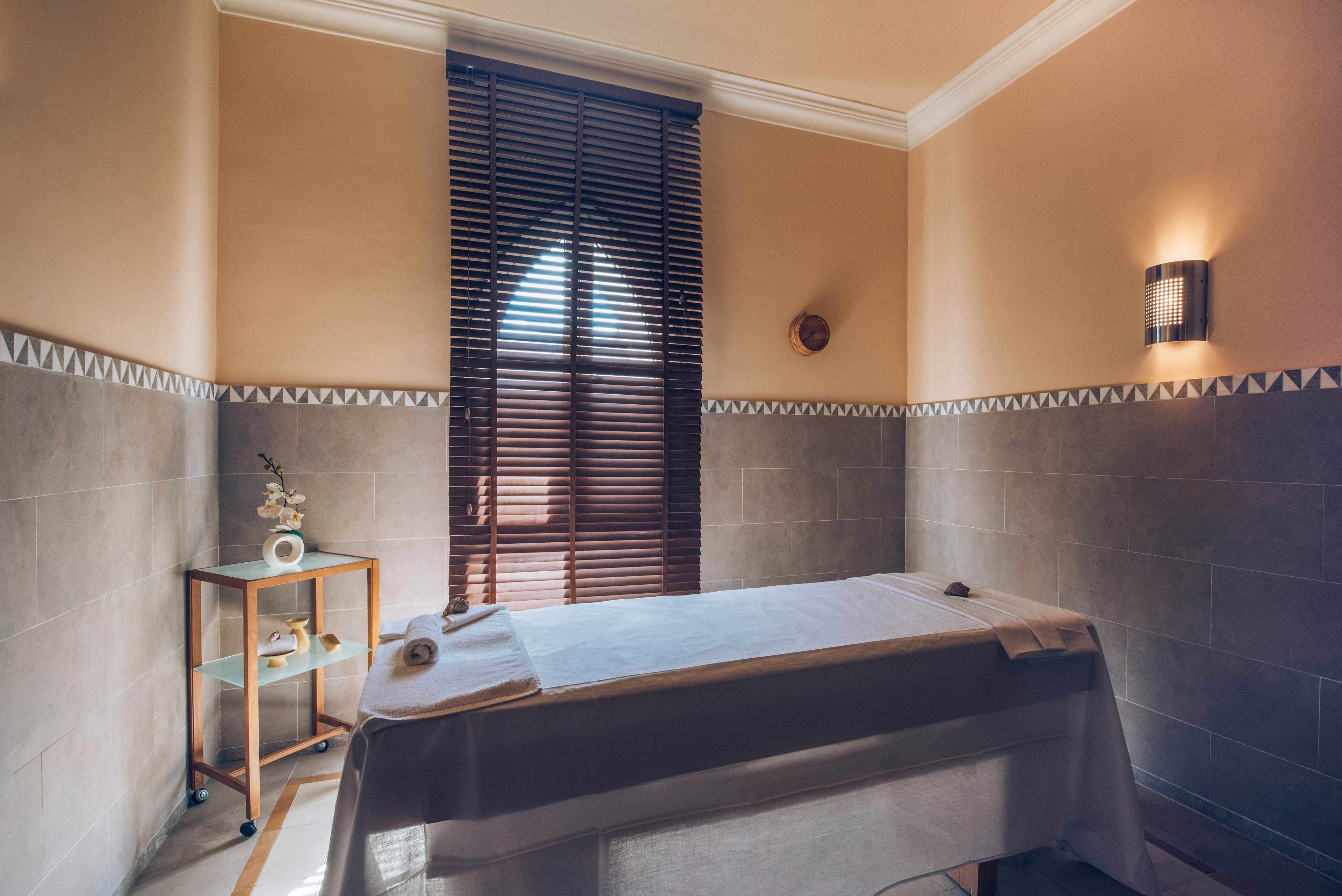 A tranquil spa treatment room features a massage table covered with crisp white linens. Warm beige and gray walls create a soothing atmosphere, complemented by soft wall sconces. Wooden blinds filter natural light from a tall window, casting gentle shadows across the space. A small side cart holds towels, candles, and decorative items, enhancing the relaxing, minimalist setting.