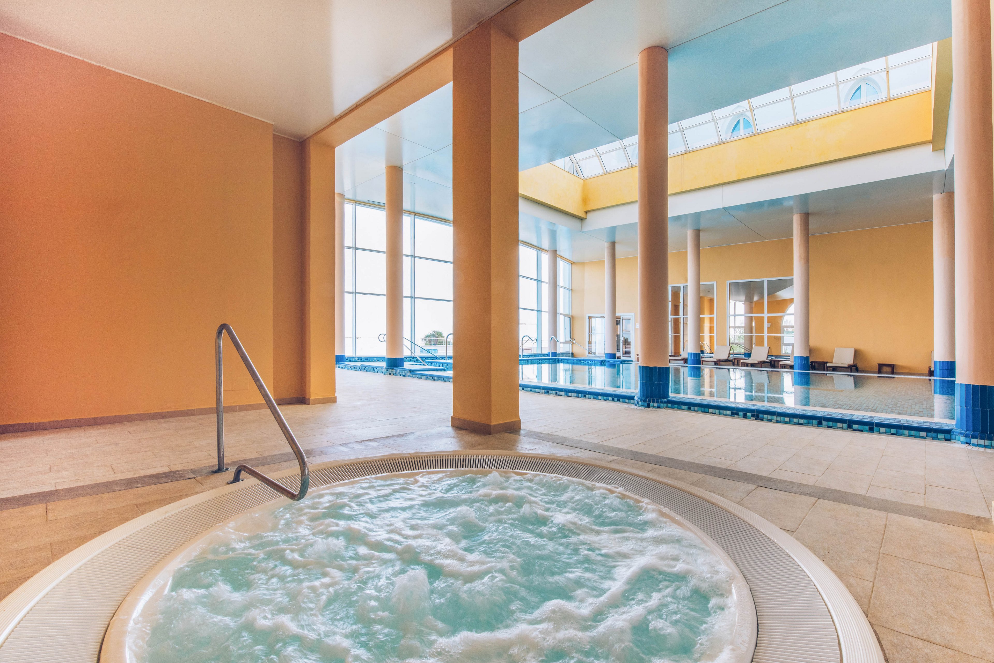 Bright indoor spa area featuring a bubbling circular jacuzzi in the foreground. A large rectangular swimming pool extends into the background, framed by tall columns and wide windows. Natural light floods the space through the glass ceiling and large wall windows, creating a calm and airy atmosphere. Warm beige walls and tiled floors contrast with the cool blue water for a relaxing wellness setting.