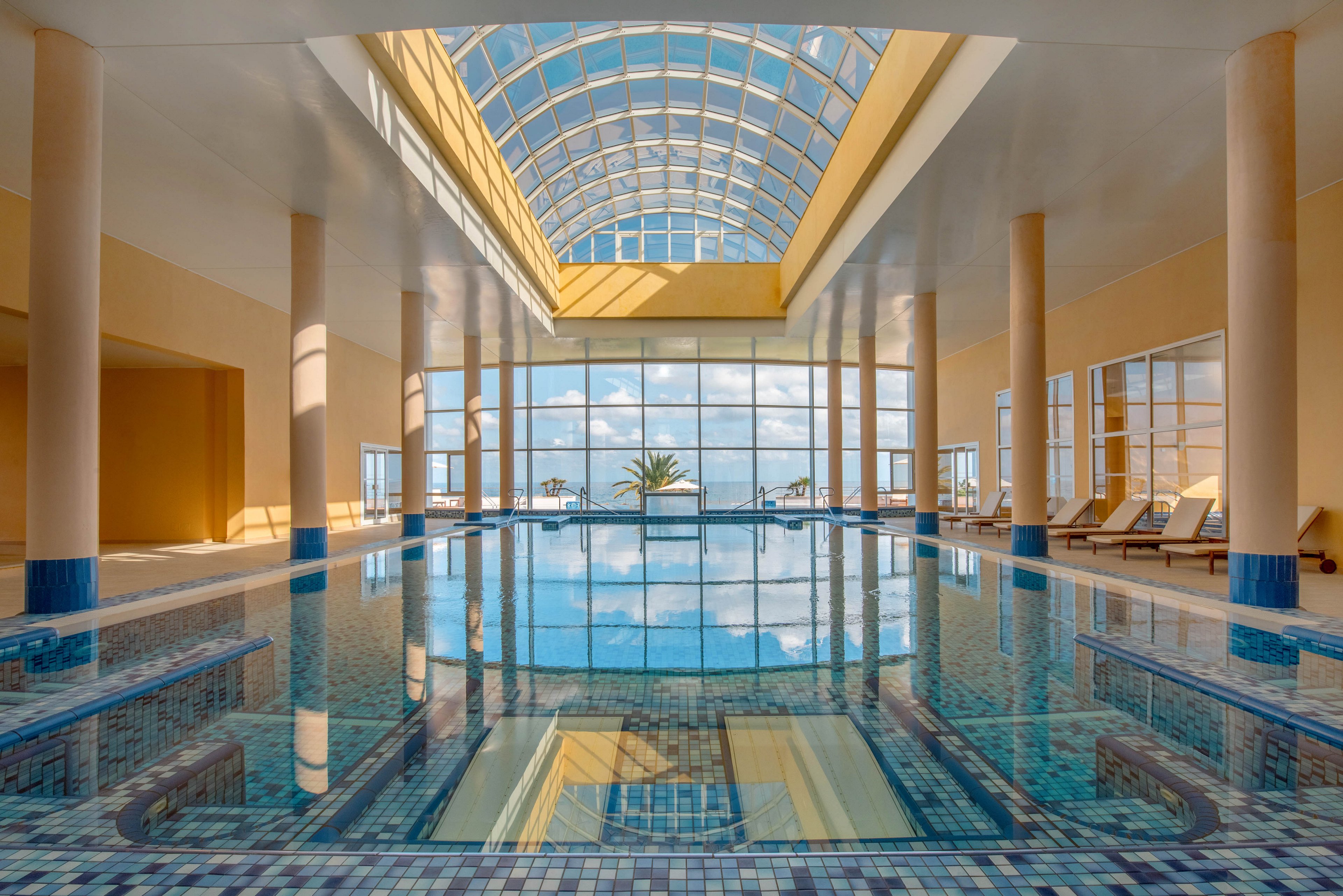 A serene indoor swimming pool stretches toward large floor-to-ceiling windows overlooking the ocean. Sunlight pours through a high arched glass ceiling, reflecting on the calm blue water and tiled floor. Lounge chairs line the sides of the pool area, suggesting a relaxing resort or spa environment. Soft beige walls and columns contrast with the cool blue tones, creating a tranquil and inviting atmosphere.