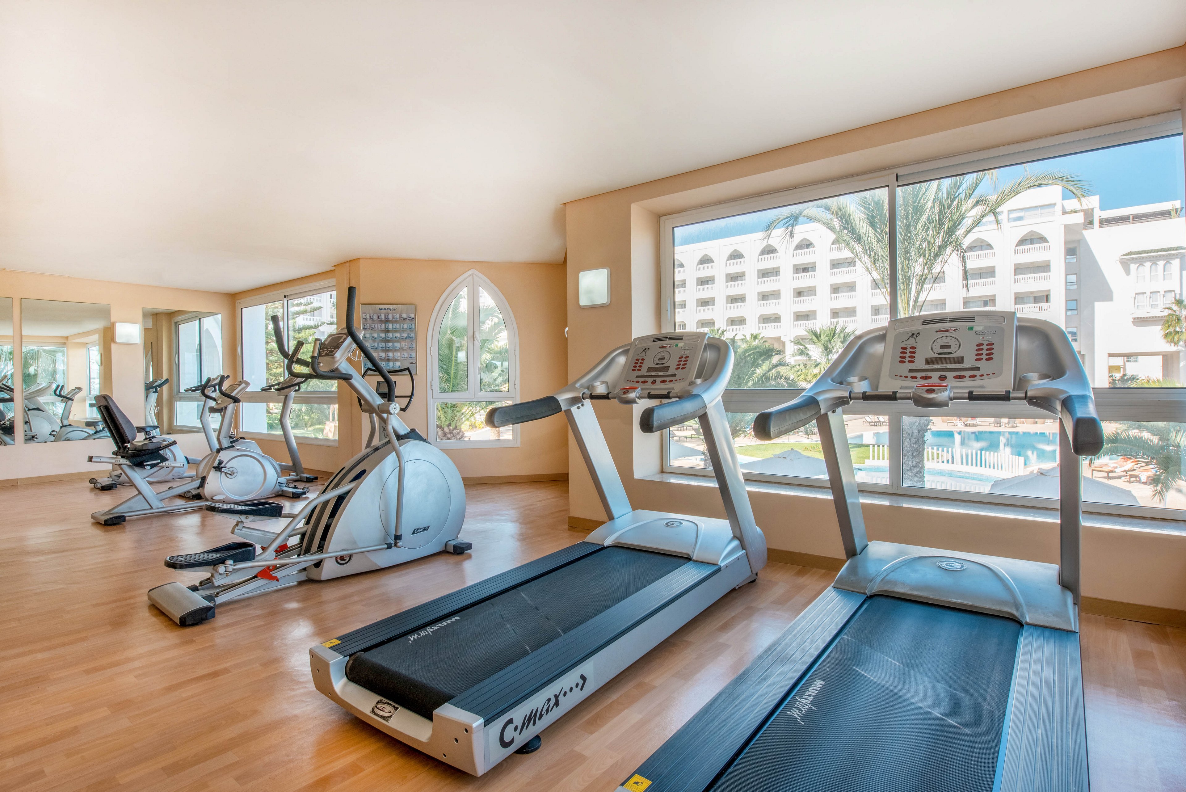 A modern hotel gym features several cardio machines, including treadmills and elliptical trainers, arranged on a polished wooden floor. Large windows provide abundant natural light and offer a view of an outdoor pool and resort buildings. Mirrors along the wall visually expand the space and reflect the exercise equipment. The room appears clean, airy, and inviting for guests to work out.