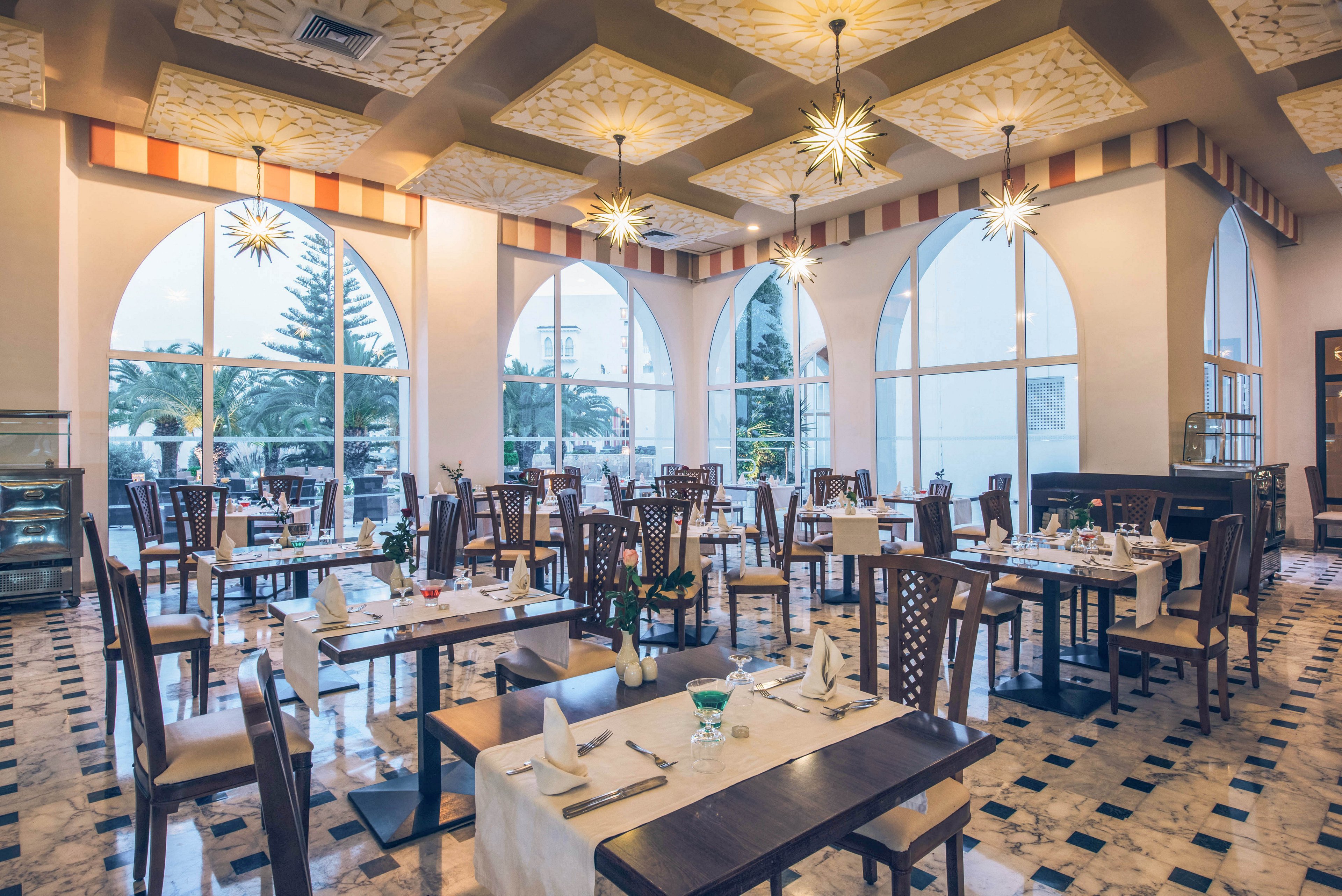 A stylish restaurant dining room is set with neatly arranged tables and chairs. Large arched windows let in soft evening light and reveal trees and buildings outside. Decorative ceiling lights and patterned ceiling panels create a warm, inviting ambiance. The tables are prepared with white tablecloths, folded napkins, glassware, and cutlery, ready for guests to arrive.