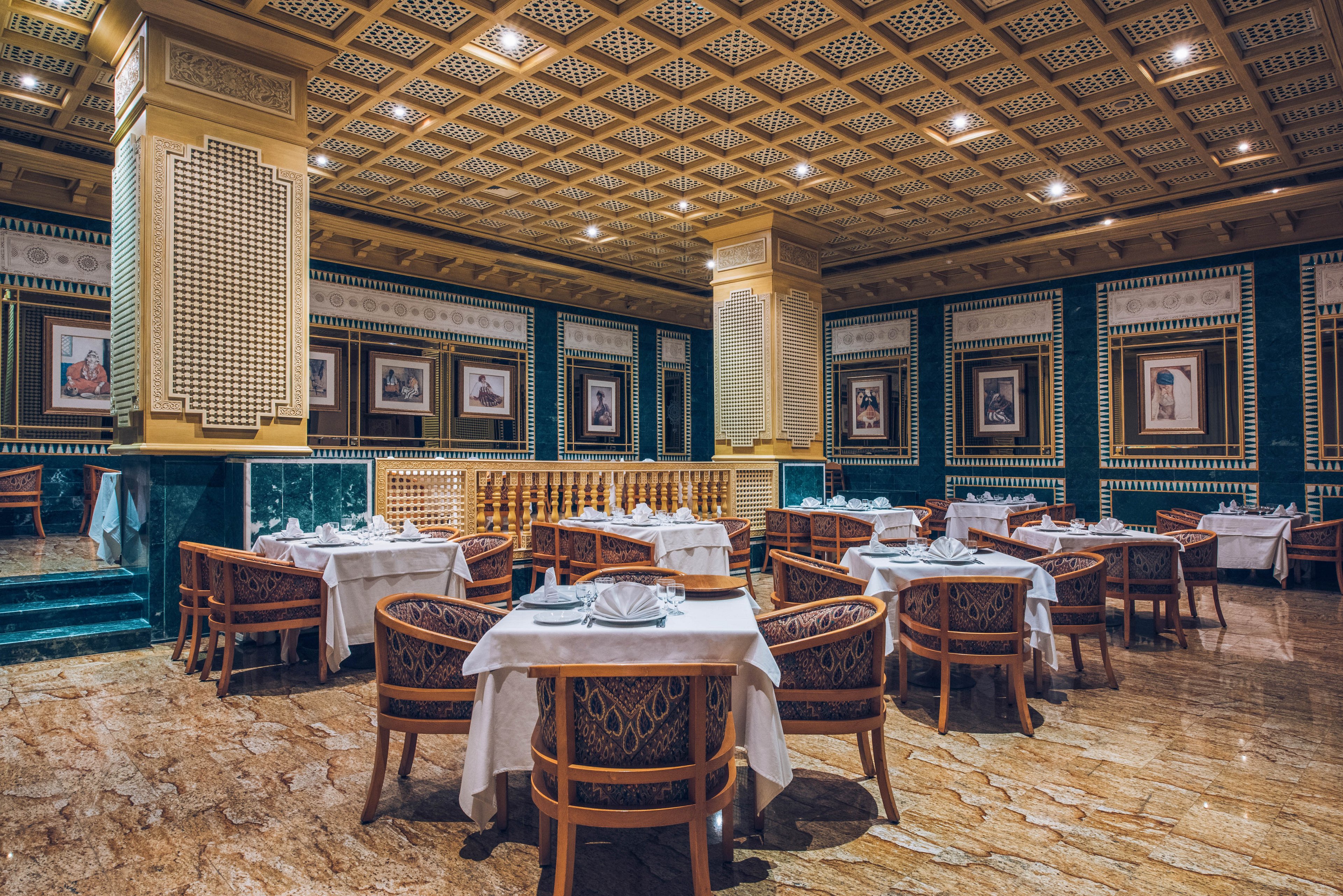 An upscale restaurant dining room features neatly arranged round tables covered with white tablecloths. Wooden chairs with cushioned seats surround each table, set with folded napkins and glassware. The space is richly decorated with patterned walls, framed artwork, and ornate ceiling panels with recessed lighting. Polished marble flooring and warm ambient light create a luxurious, formal atmosphere.