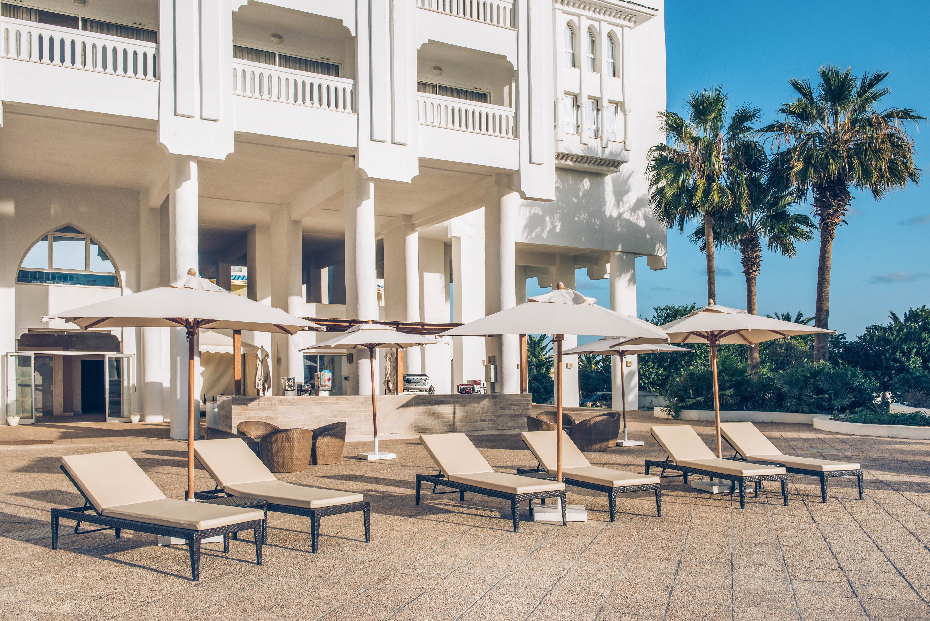 A row of beige sun loungers with umbrellas is arranged on a spacious terrace in front of a grand white resort building. The scene is set outdoors on a sunny day with clear blue skies. Tall palm trees frame the area, adding a tropical feel to the modern architecture. The overall atmosphere suggests a relaxing vacation setting by the sea or pool.
