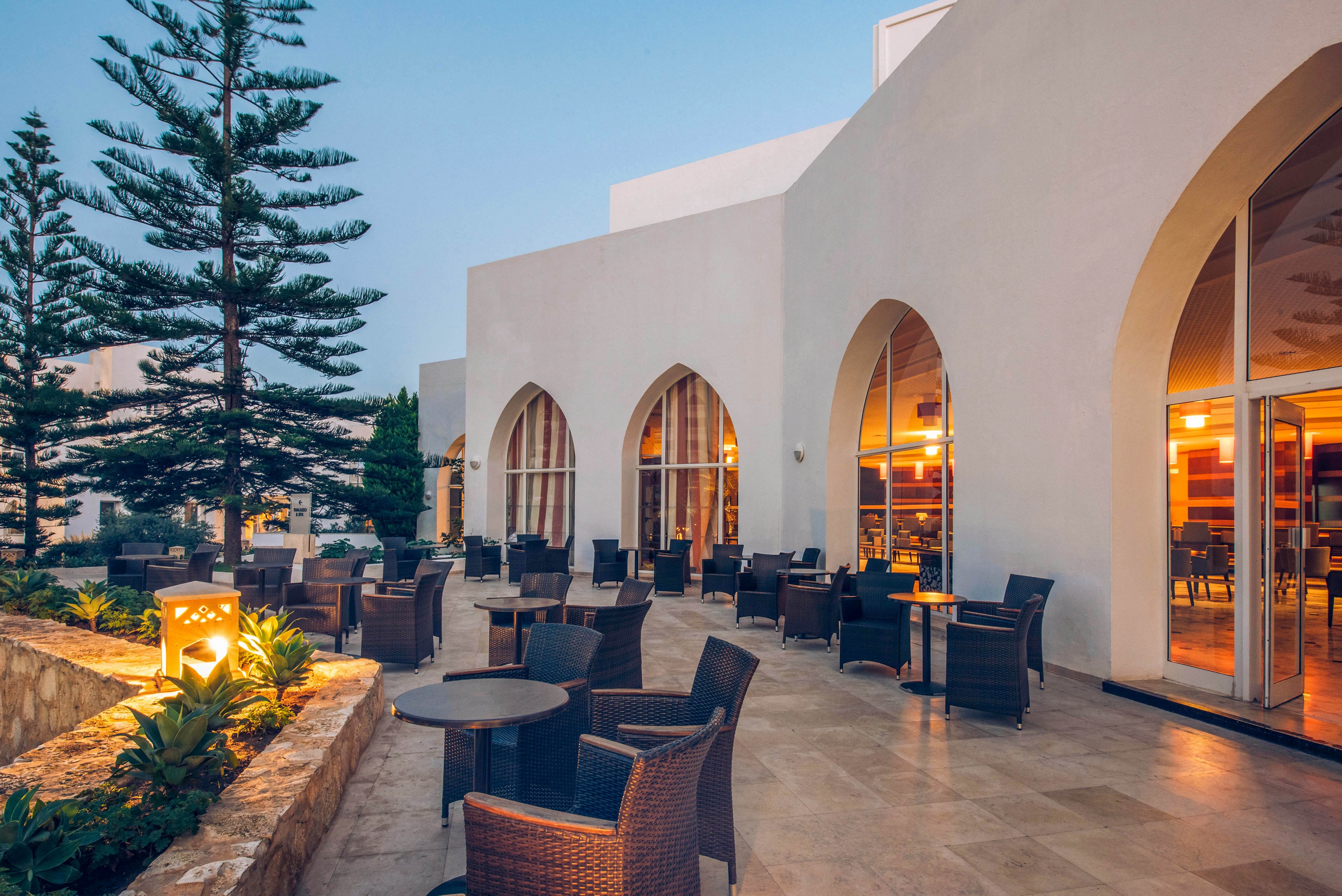 An outdoor hotel terrace features wicker chairs and round tables arranged on a spacious tiled patio. The white building has large arched windows glowing warmly from interior lights. Tall evergreen trees and low stone planters with greenery frame the seating area. The scene is captured at dusk under a soft blue sky, creating a calm and inviting atmosphere.
