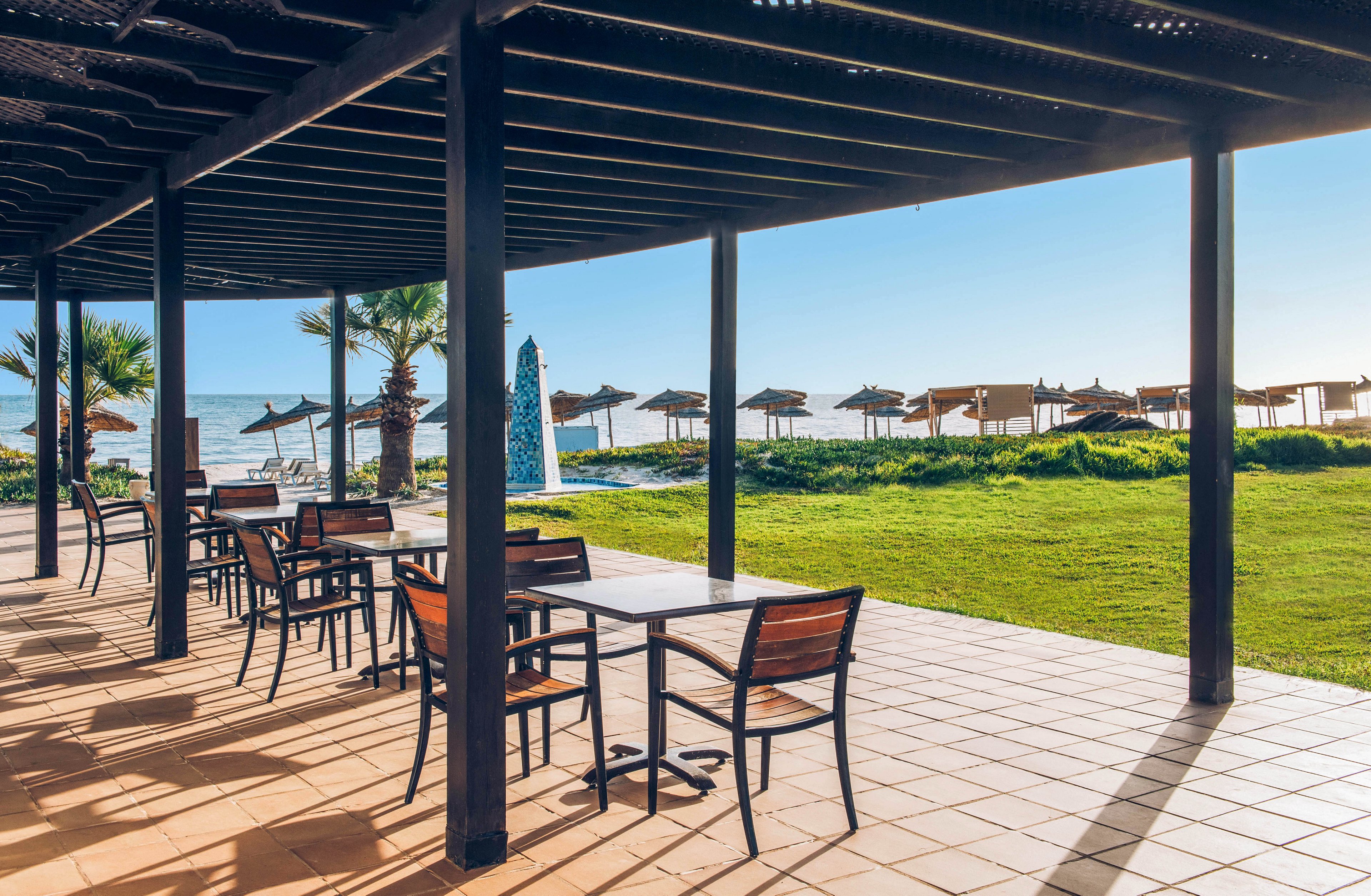 Open-air terrace seating overlooks a sunny beachfront with rows of straw umbrellas. Empty wooden tables and chairs sit under a shaded pergola on a tiled patio. A green lawn separates the terrace from the sandy shore and calm blue sea. The clear sky and bright light create a relaxed, vacation atmosphere.