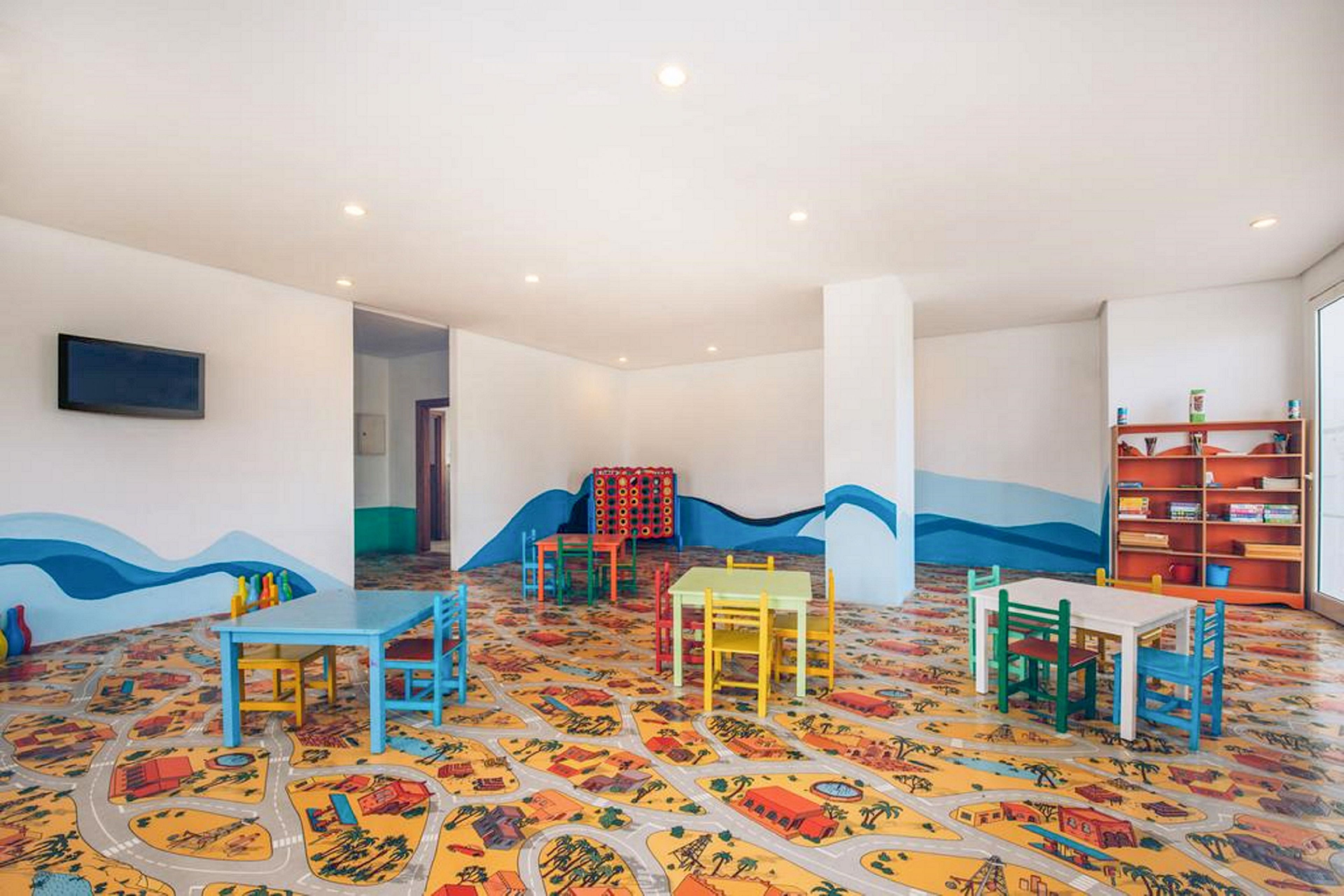 A spacious children’s playroom features several small colorful tables and chairs arranged across a vibrant patterned floor. The floor design shows playful town and road illustrations, adding a whimsical feel to the space. White walls with blue wave graphics surround the room, and a flat-screen television is mounted on one wall. Shelving with toys and games is visible in the background, suggesting an inviting area for kids’ activities.