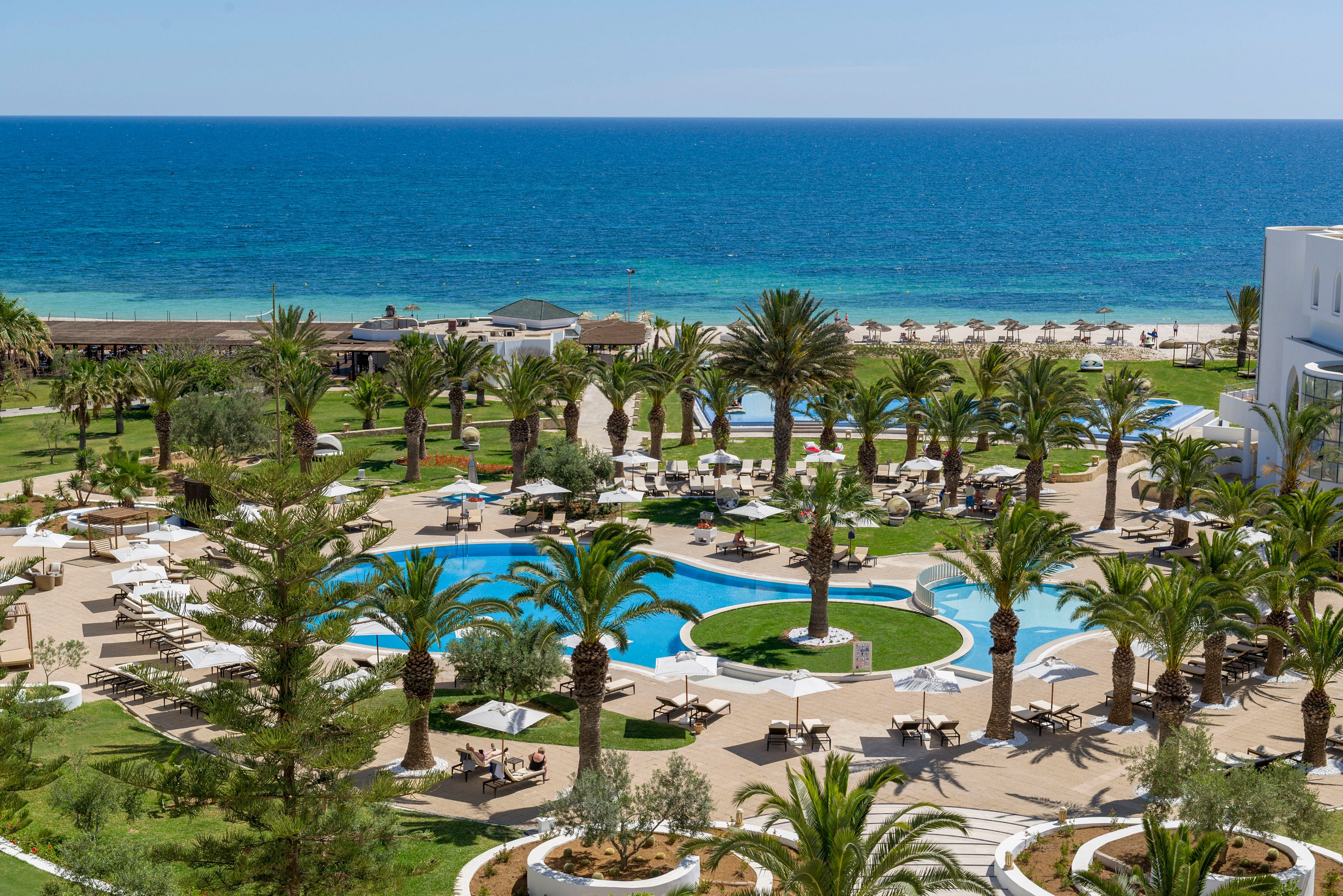 A large seaside resort features multiple outdoor swimming pools surrounded by palm trees and sun loungers. White buildings frame the landscaped gardens, creating a bright and inviting vacation setting. Guests relax under umbrellas near the pools, with pathways winding through the green lawns. In the background, a wide sandy beach meets the deep blue ocean under a clear sky.