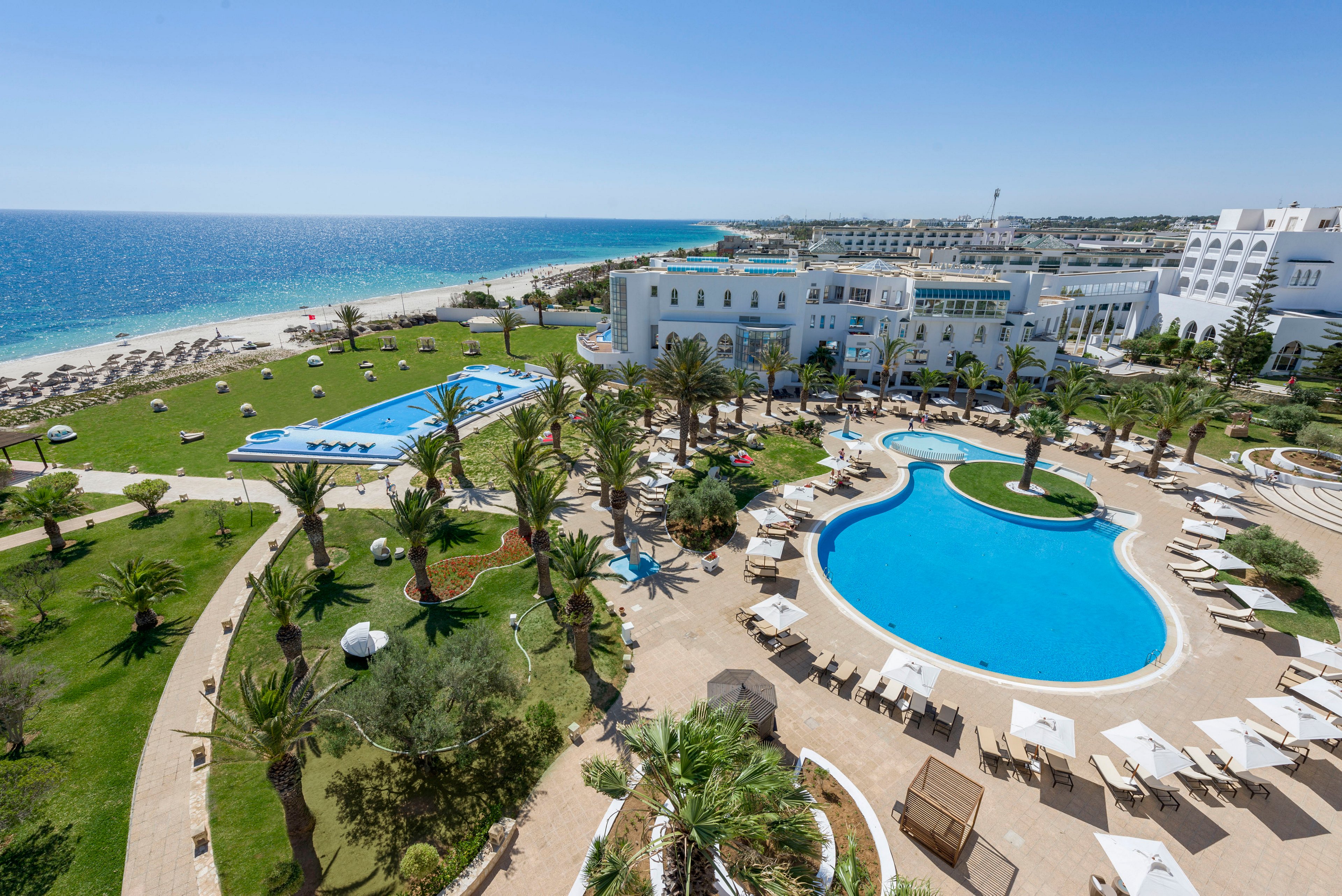 A large coastal resort features two outdoor swimming pools surrounded by palm trees and sun loungers. White buildings with arched windows overlook the landscaped gardens and pathways. The property extends to a sandy beach lined with umbrellas and chairs beside a calm blue sea. The bright daylight and clear sky create a relaxing vacation atmosphere.
