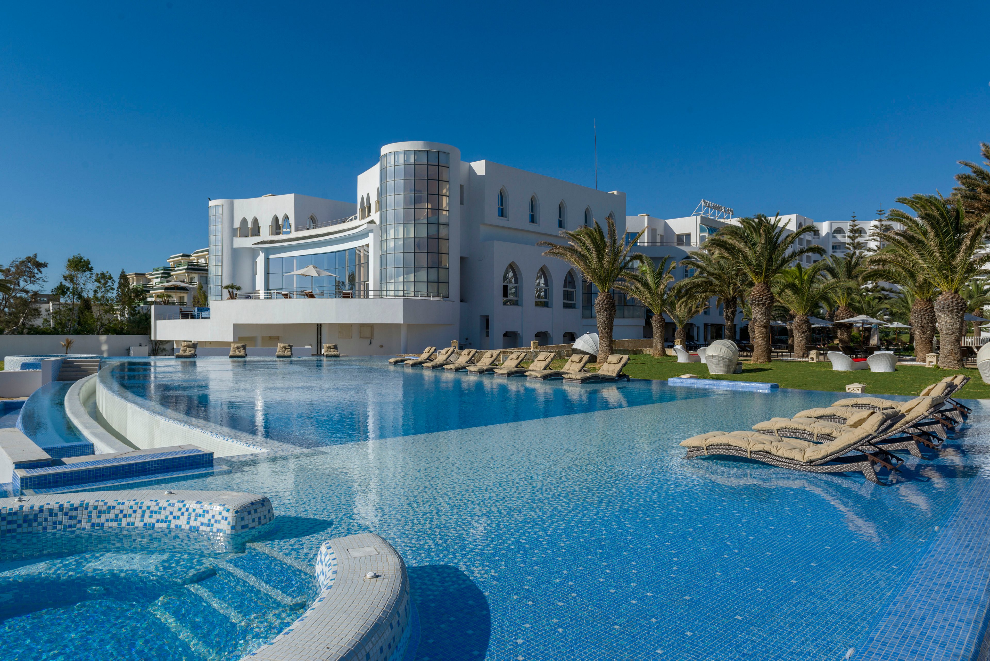 A large, modern white resort building overlooks an expansive outdoor swimming pool under a clear blue sky. Numerous sun loungers line the poolside, suggesting a relaxing vacation setting. Palm trees and manicured lawns add a tropical atmosphere to the scene. The calm water and bright sunlight create a serene and inviting environment.