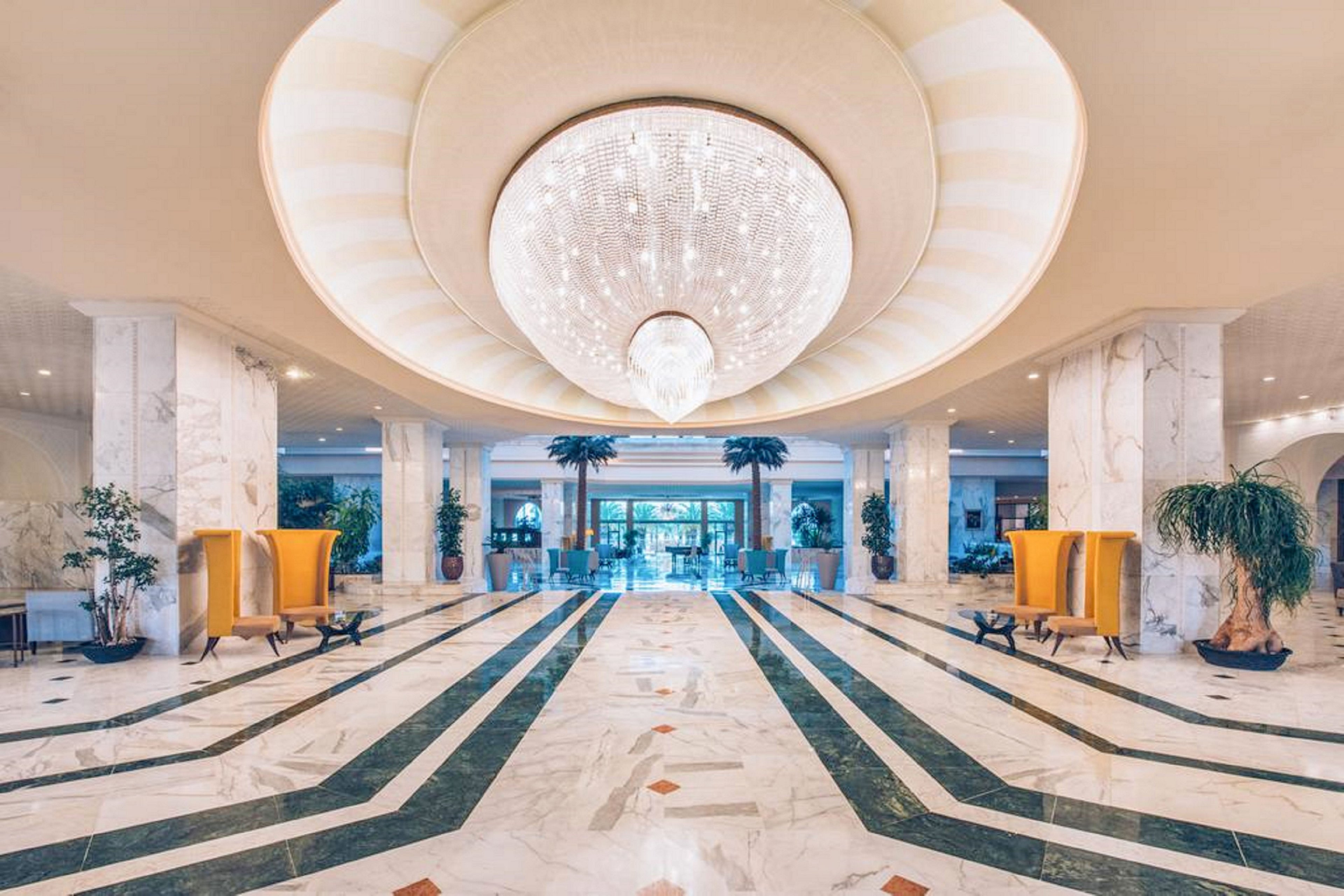 Spacious, marble-clad hotel lobby featuring a large crystal chandelier as the central focal point. Symmetrical green and white floor patterns lead toward glass entrance doors and an outdoor view. Tall indoor plants and modern yellow seating alcoves line both sides of the hall, adding color and texture. The bright, well-lit interior conveys an elegant, upscale atmosphere suitable for hospitality and events.