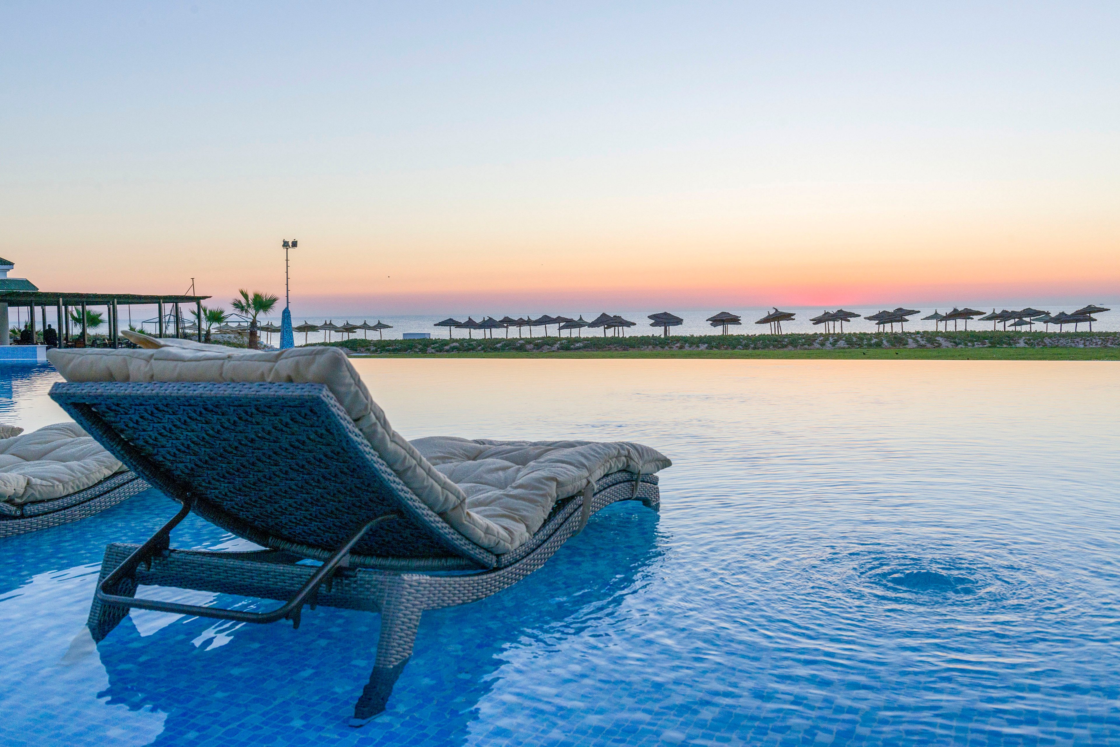 Two cushioned wicker loungers sit partially submerged in a tranquil infinity pool, facing a calm sea at sunset. The horizon glows with soft pastel colors as the sky transitions from blue to warm orange and pink. In the distance, a row of beach umbrellas and a low resort structure line the waterfront, suggesting a luxurious vacation setting. The overall scene conveys relaxation, leisure, and peaceful coastal ambiance.