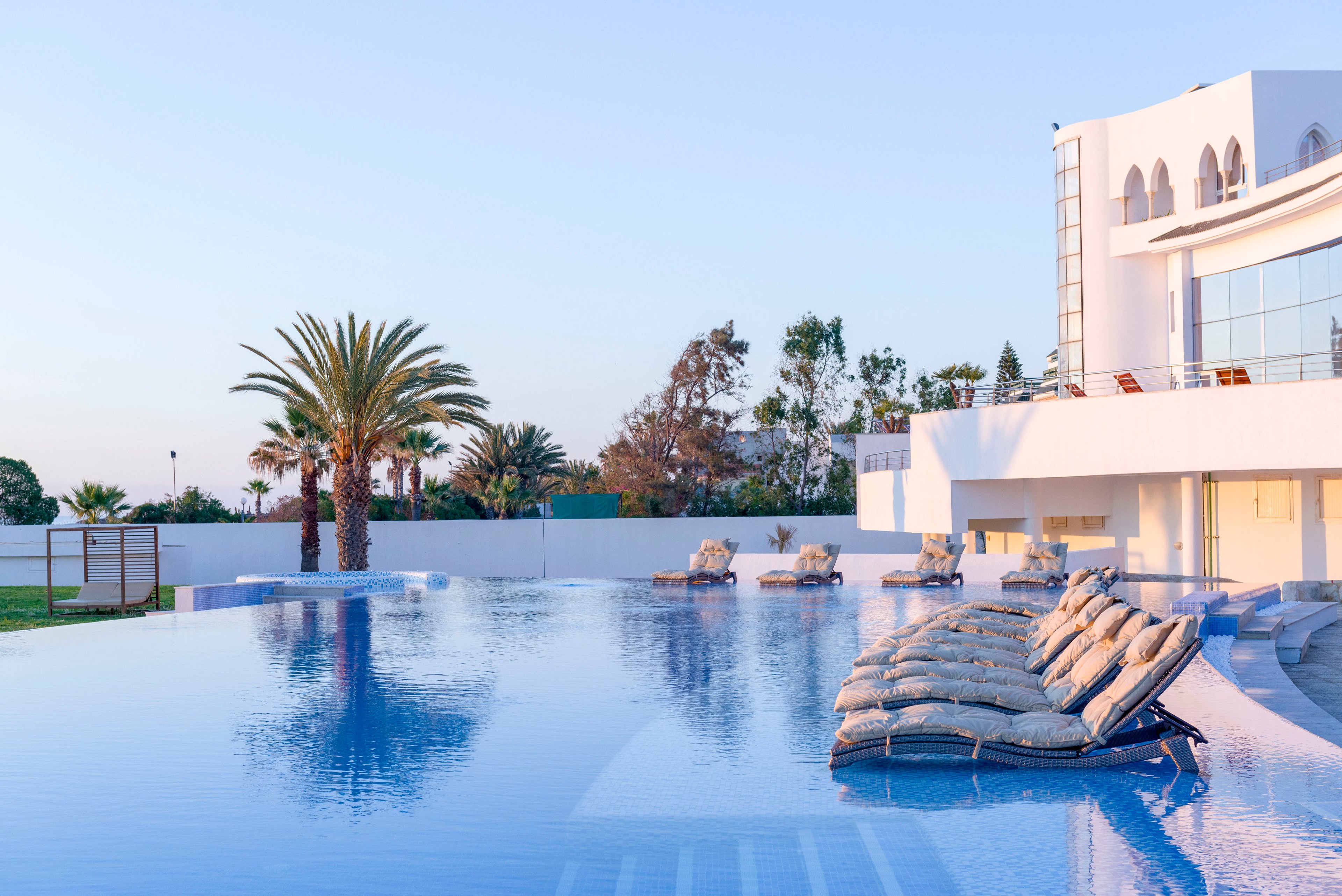 A large infinity pool stretches out beside a modern white villa at sunset. Cushioned lounge chairs line the poolside, facing the calm water and distant horizon. Palm trees and landscaped greenery frame the property, adding a tropical touch to the serene setting. Soft pastel sky tones reflect on the water, creating a tranquil and luxurious atmosphere.