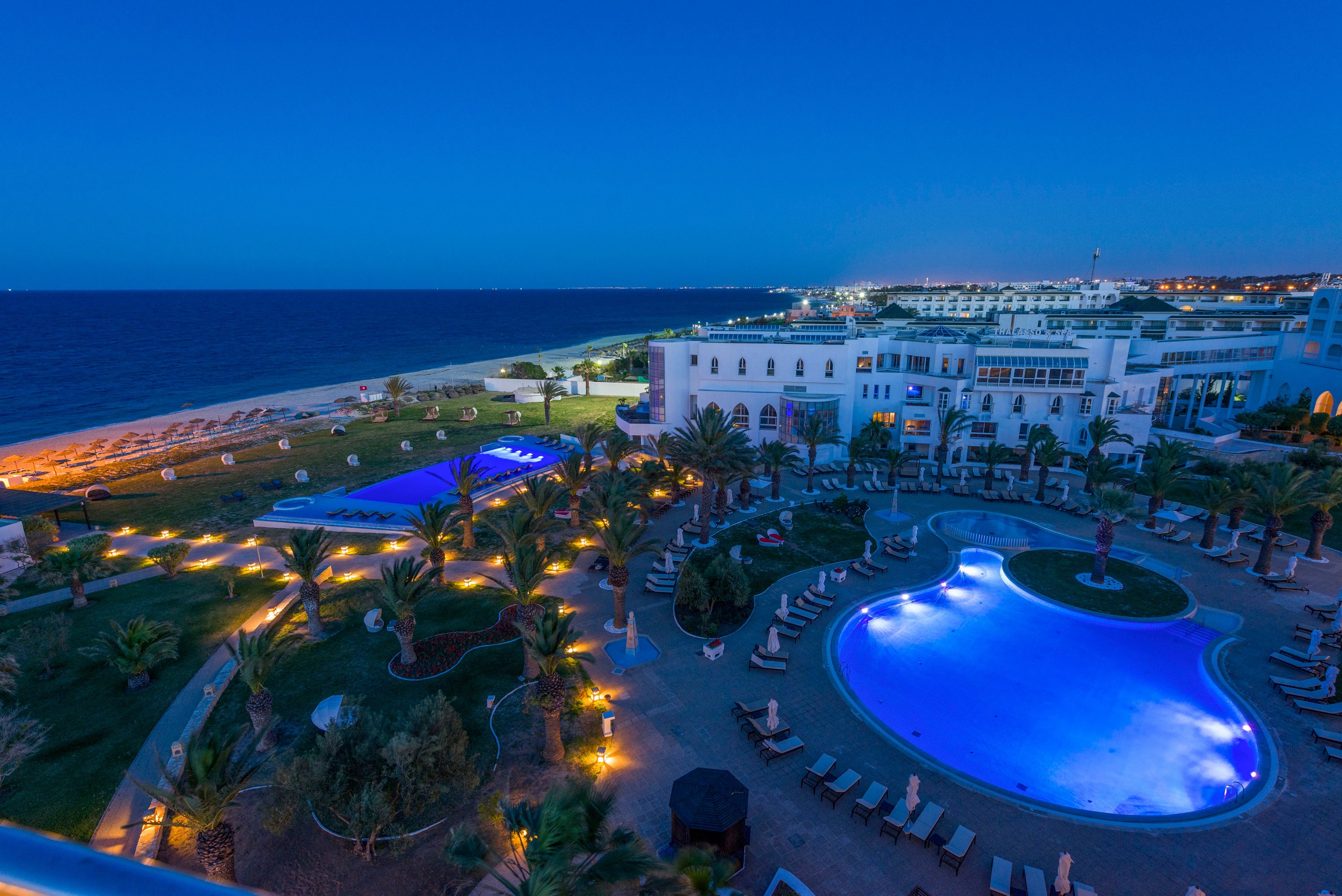 A large seaside resort is shown at dusk with two brightly lit swimming pools surrounded by lounge chairs and palm trees. The hotel buildings are positioned near the shoreline, overlooking a calm, deep blue sea. Warm pathway lights and pool lighting create a striking contrast against the cool evening sky. The expansive lawn and landscaped gardens emphasize the relaxing vacation atmosphere.