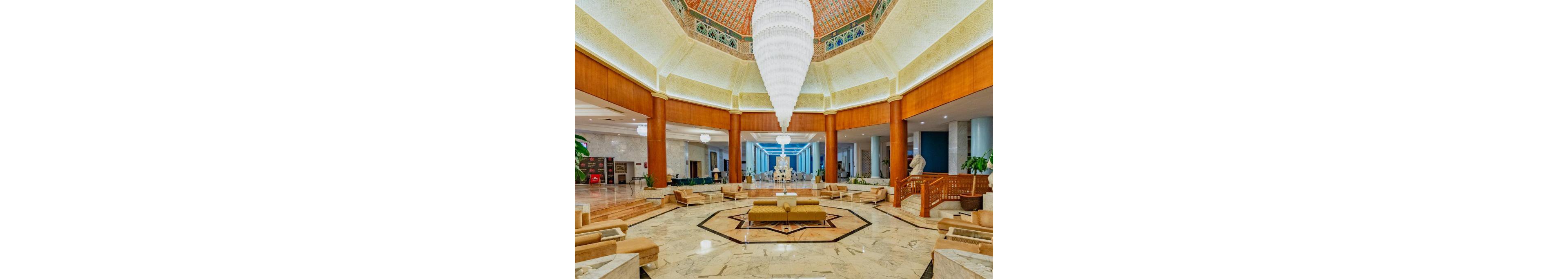 Spacious hotel lobby interior featuring a dramatic central chandelier and marble flooring. Symmetrical seating areas with beige sofas and armchairs surround a star-shaped inlay on the floor. Tall wooden columns frame the space, leading the eye toward a statue and corridor in the background. Warm lighting and neutral tones create an elegant, welcoming atmosphere suitable for upscale hospitality settings.