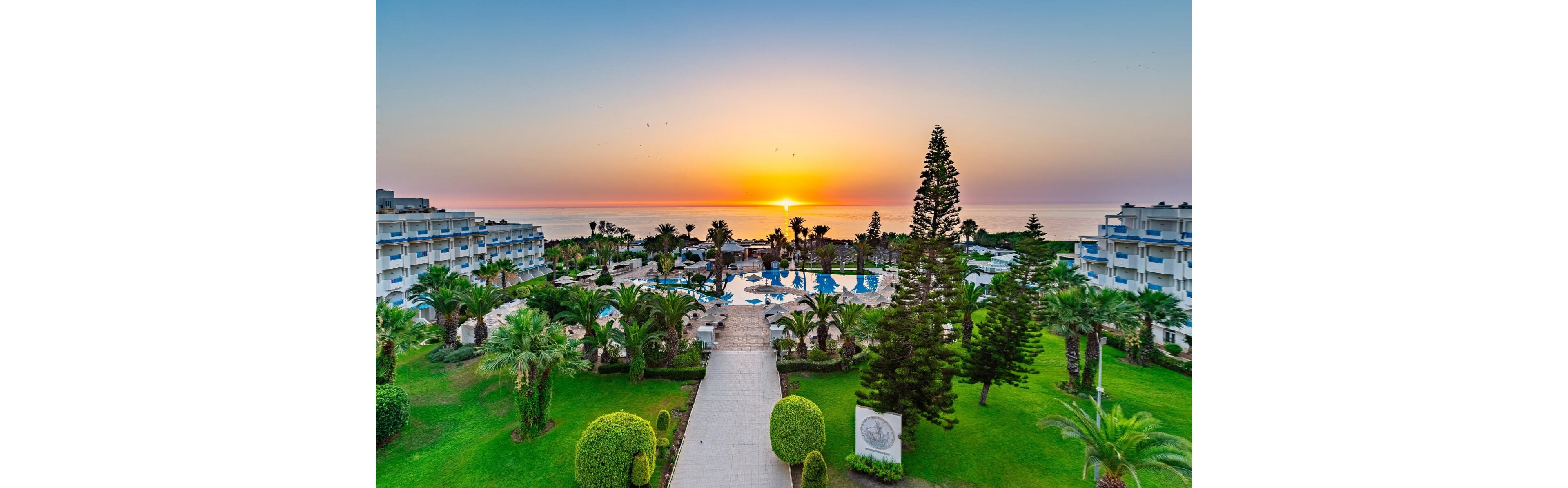 A large seaside resort complex is shown at sunset, with the sun setting over the ocean in the distance. In the foreground, a straight pathway leads through manicured lawns and lush palm trees toward multiple swimming pools. White multi-story hotel buildings frame both sides of the scene, creating a symmetrical composition. The sky glows with warm orange and pink tones, reflecting softly on the water and enhancing the tranquil vacation atmosphere.