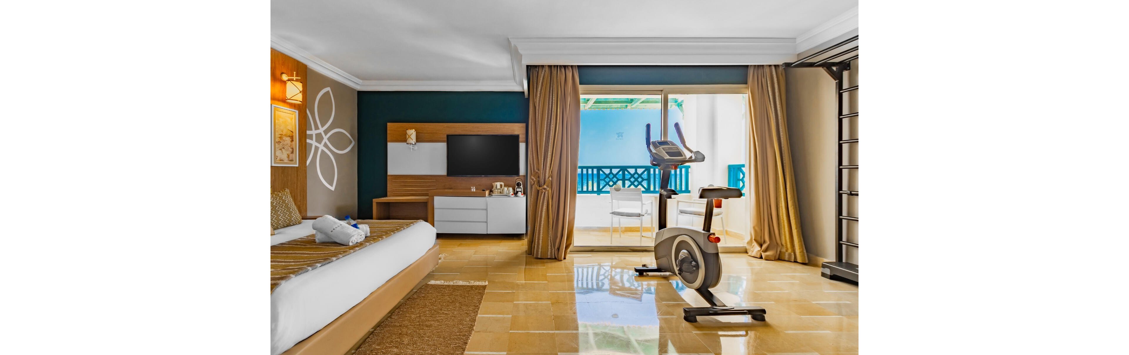A spacious modern hotel room features a neatly made bed and a large flat-screen television mounted on a wooden media unit. An exercise bike is positioned near the sliding glass doors that open to a bright balcony with blue railings. Warm tan floor tiles and golden curtains create a cozy, inviting atmosphere. Decorative wall lighting and simple artwork add a touch of contemporary style to the interior.