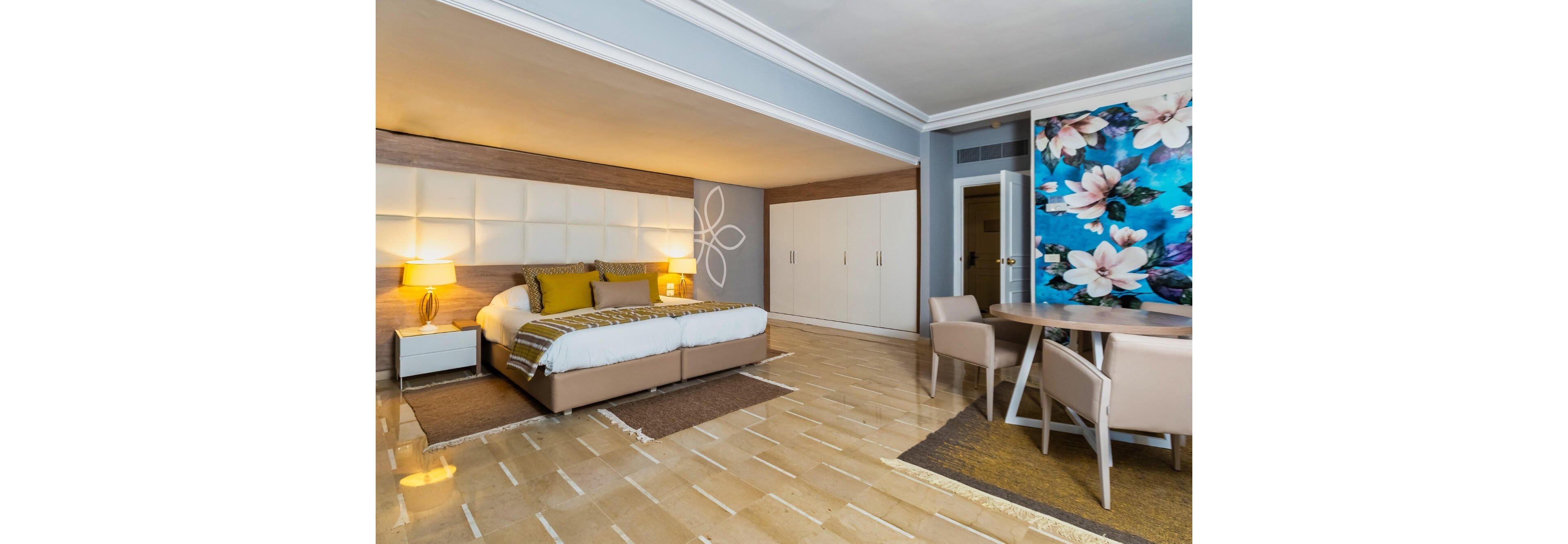 Spacious contemporary hotel room featuring a large double bed with white linens and mustard accent pillows. The setting includes bedside tables with warm lamps, a built-in white wardrobe, and a small dining or work table with chairs. A striking floral mural in blue and pink tones decorates one wall, adding color to the neutral interior. Polished tile flooring and soft area rugs complete the clean, modern aesthetic.