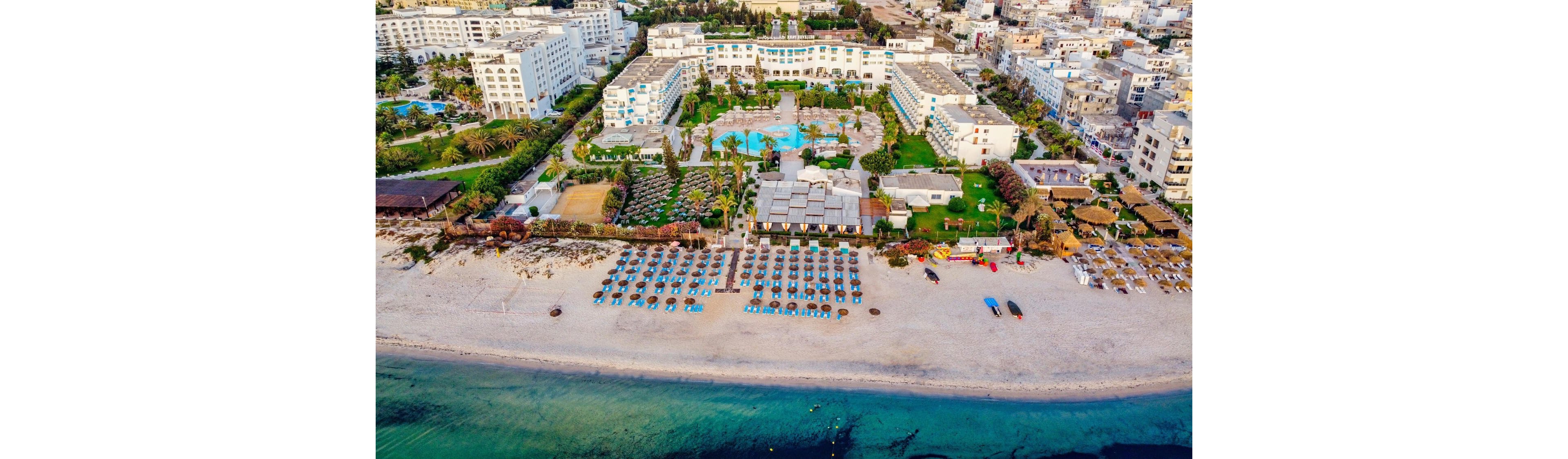 An aerial view shows a large beachfront resort complex facing a sandy shoreline. Rows of sun loungers and umbrellas are neatly arranged on the beach beside calm turquoise water. Behind the beach, multiple swimming pools, gardens, and white hotel buildings are visible. The resort is surrounded by a dense urban area with numerous low and mid-rise buildings.