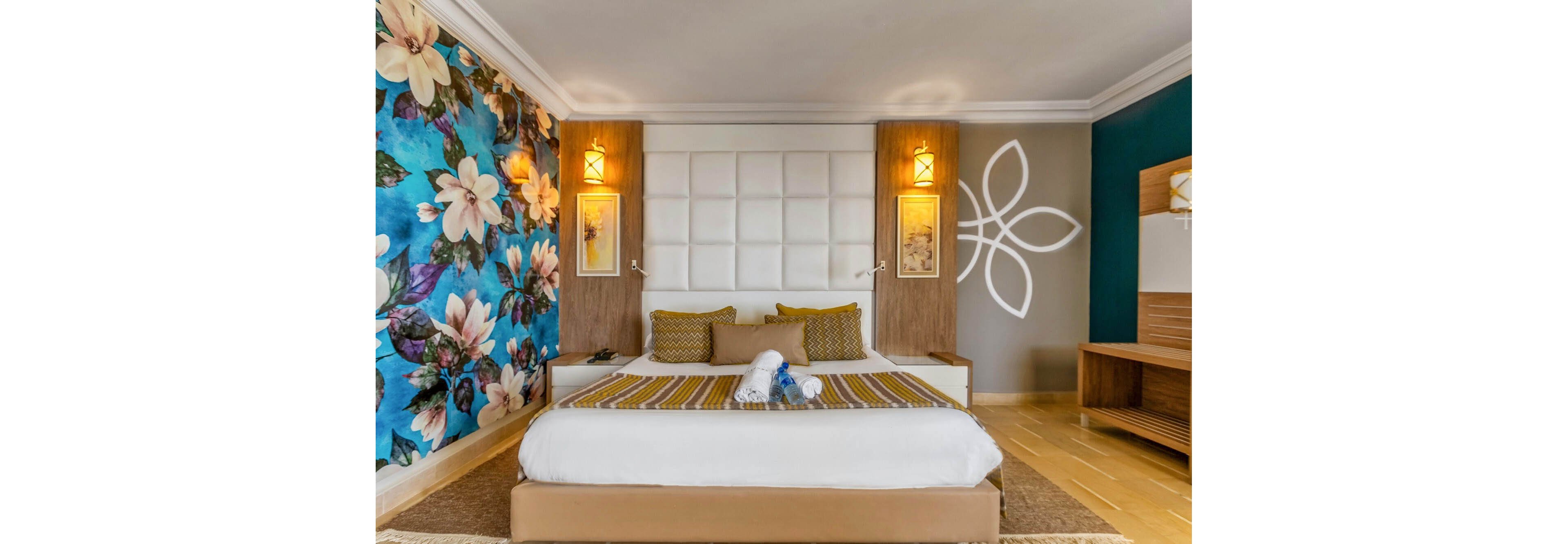 A neatly made double bed sits at the center of a modern hotel bedroom. The room features a bold floral accent wall on one side and a large white padded headboard framed by wooden panels. Warm pendant lights, decorative wall art, and folded towels shaped like swans add a welcoming touch. A desk and shelving unit are visible to the right, completing the contemporary, cozy atmosphere.