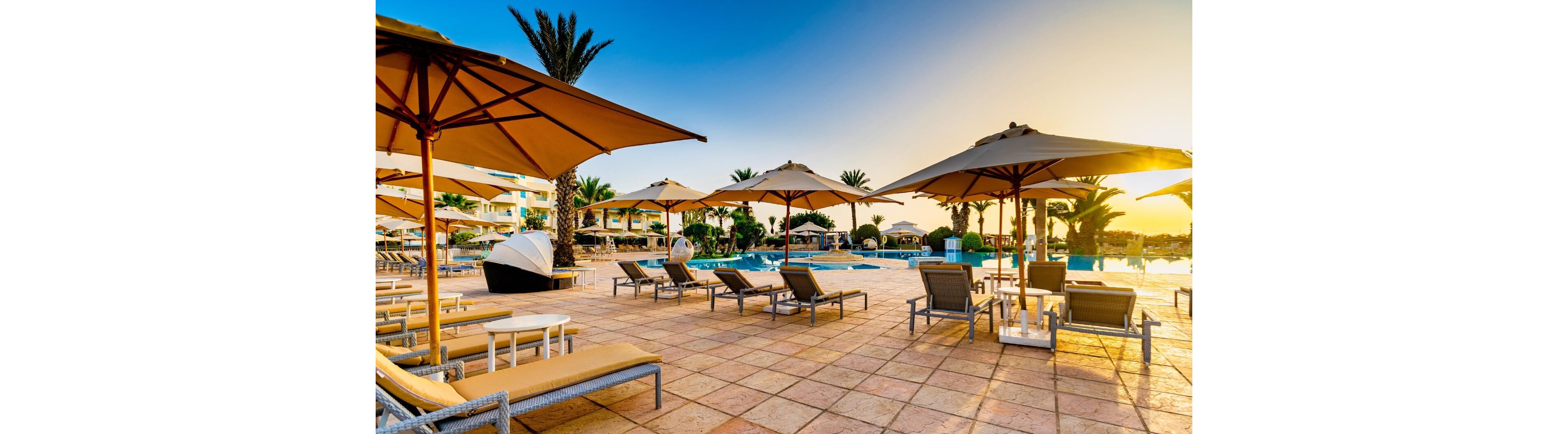Outdoor lounge chairs and umbrellas surround a tranquil resort swimming pool at sunset. The scene features tiled flooring, palm trees, and calm blue water reflecting the warm sky. Empty sunbeds and shaded seating areas suggest a peaceful, relaxing vacation atmosphere. The golden light enhances the inviting, tropical feel of the poolside setting.