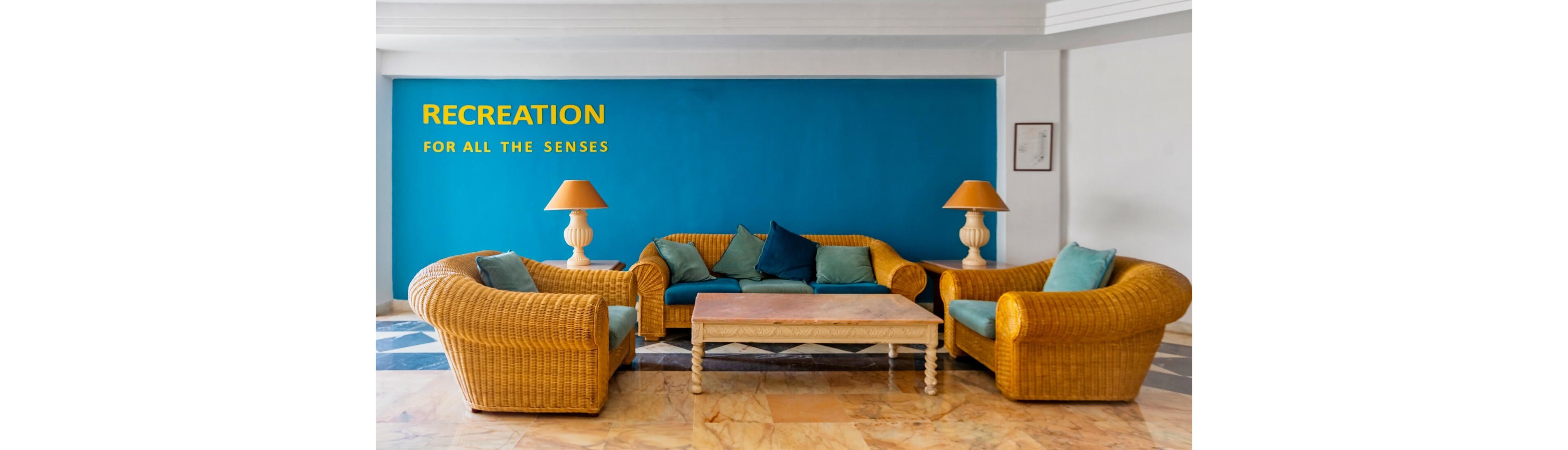 A set of wicker sofas and armchairs with teal and blue cushions is arranged around a wooden coffee table in a bright indoor lounge. The seating area is placed against a bold turquoise wall that features the text "RECREATION FOR ALL THE SENSES" in yellow. Two matching table lamps with orange shades sit on either side of the central sofa. The floor has a mix of marble and patterned tiles, giving the space a relaxed yet stylish resort ambiance.