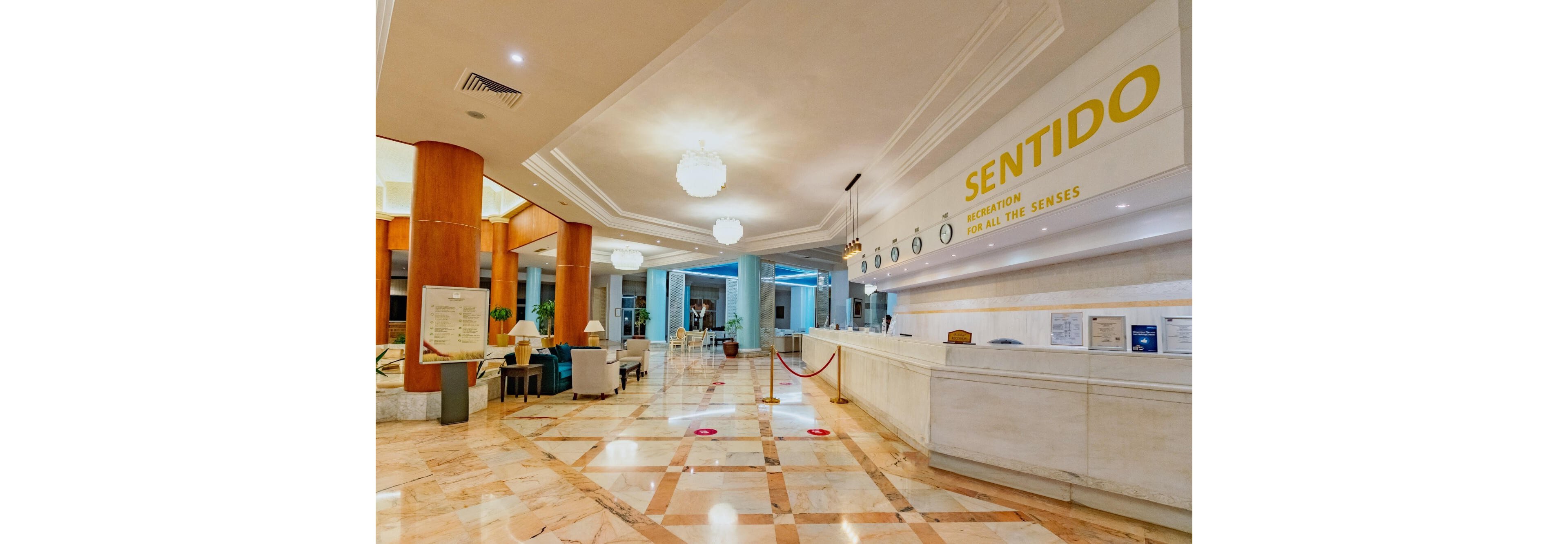 A bright, spacious hotel lobby features a long white reception desk with the word SENTIDO prominently displayed on the wall. The polished marble floor reflects warm ceiling lights and circular chandeliers. Seating areas with armchairs and tables are visible to the left, while a roped-off queue guides guests at the front desk. The atmosphere feels modern, clean, and welcoming, with soft yellow and blue tones throughout the space.
